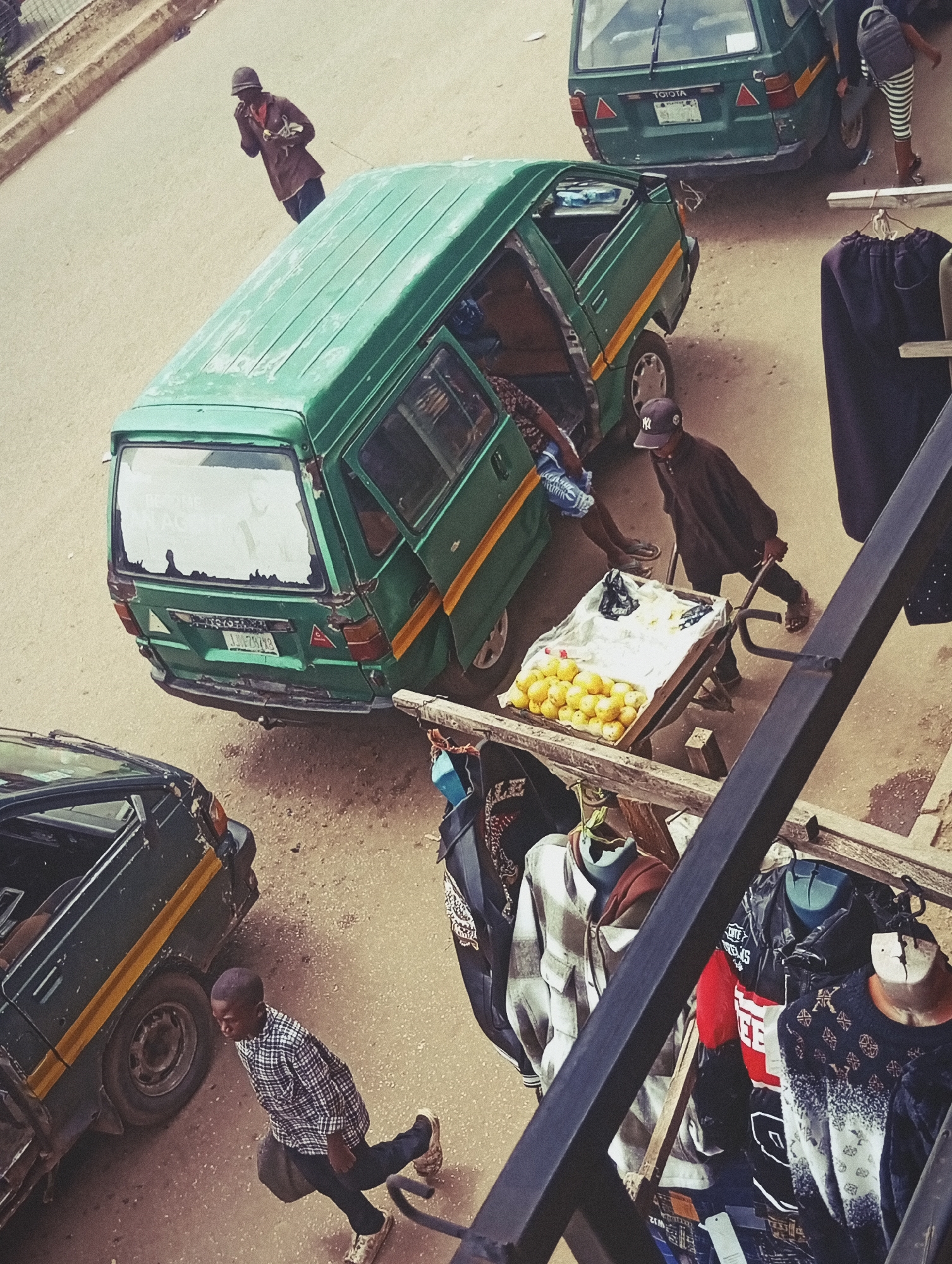 The image depicts a bustling scene in Bukuru, Nigeria, likely during daylight hours given the shadows. The primary focus is on a street-side market activity centered around two green minibuses, commonly used for public transportation in the region.
In the foreground, a young boy in a checkered shirt walks with a bag slung over his shoulder, his gaze directed downwards. He appears to be navigating the busy street, possibly assisting with the market or on his way to or from it. Behind him, a green minibus is parked, its doors open, suggesting it might be stopped for passengers or goods.
Further into the scene, another green minib us is the main subject. Its back is towards the viewer, and it has the "TOYOTA" logo visible on the rear. A person wearing a dark outfit and a backward baseball cap is seen pushing a cart laden with bright yellow fruits, likely oranges or tangerines, which are neatly arranged in a wooden crate. This suggests a vendor is actively selling produce. Another person, partially obscured, appears to be inside or near the open door of this minibus.
In the background, a figure in a brown shirt and a hat is seen walking away, holding something small in their hands. The ground is a sandy, dusty road, typical of many Nigerian urban settings. Clothing items are displayed on a makeshift rack in the right portion of the image, indicating a small roadside stall. The overall atmosphere seems to be one of commerce and daily life in a busy marketplace. No specific weather conditions are discernible, but the light suggests it is not nighttime.
