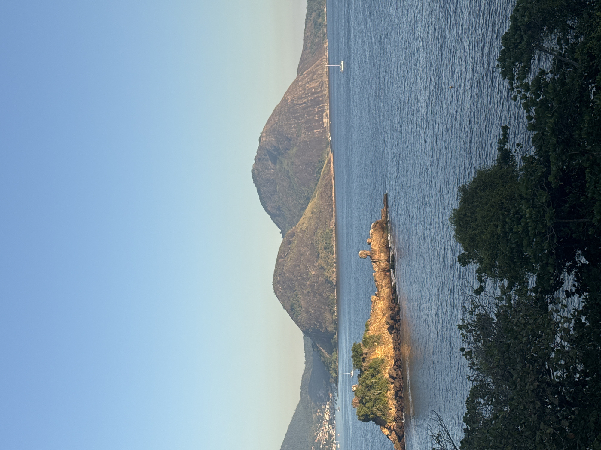 This is a scenic view of the Guanabara Bay, likely taken from Niterói, Brazil, looking towards Rio de Janeiro. In the foreground, lush green trees frame the bottom of the image. The middle ground features a small, rocky island covered with vegetation, illuminated by the sun. Several sailboats are visible on the calm blue water. In the background, two prominent mountains dominate the landscape, with a town nestled on the slopes of one of them. The sky is a clear, pale blue.