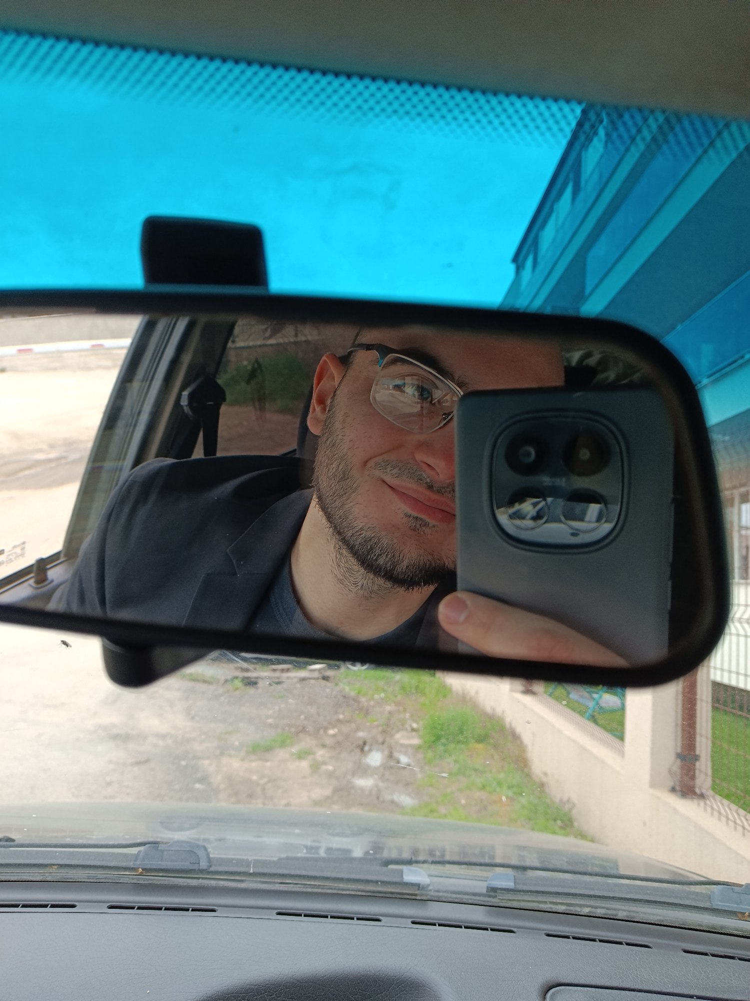 A man is taking a selfie in the rearview mirror of a car. The man has short dark hair and a beard. He is wearing glasses and a dark jacket. He is holding a dark-colored smartphone with a distinctive circular camera array. The car's windshield is visibly dirty, with the dashboard appearing dark gray. Outside the car, there is a road, some greenery, and a building with a blue facade and white columns. Based on the architecture and landscaping style, the location is likely in Plovdiv, Bulgaria.