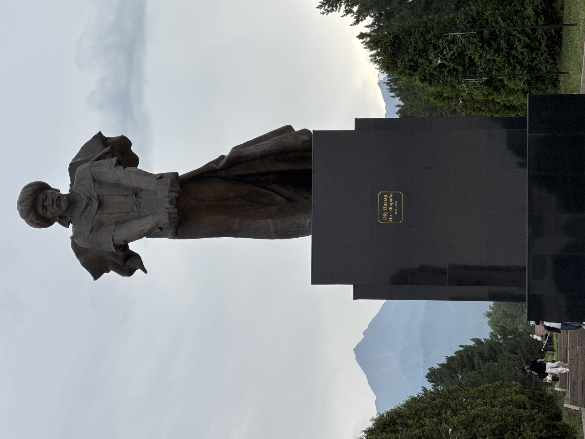 A full-shot, eye-level, outdoor photograph captures a bronze statue of a man wearing a turban and robes, standing on a tall, black granite pedestal. The statue depicts the man with his hands clasped in front of him, his gaze directed forward. The pedestal features a golden plaque with text in Cyrillic script and Arabic numerals, likely identifying the person depicted and their lifespan.
In the background, a range of mountains, possibly the Zailiysky Alatau, forms a backdrop against a hazy, overcast sky. Green conifer trees line the left and right sides of the scene, with a pathway and stairs leading up to the monument. Two individuals are visible on the stairs in the lower left quadrant, one walking down and talking on a mobile phone, the other walking down. A person in a white top and light-colored pants is also visible further down the stairs, as is another person in a dark top and jeans near the bottom. The overall impression is one of a public monument in a park or square setting, likely in Almaty, Kazakhstan, given the presence of the Zailiysky Alatau mountains.