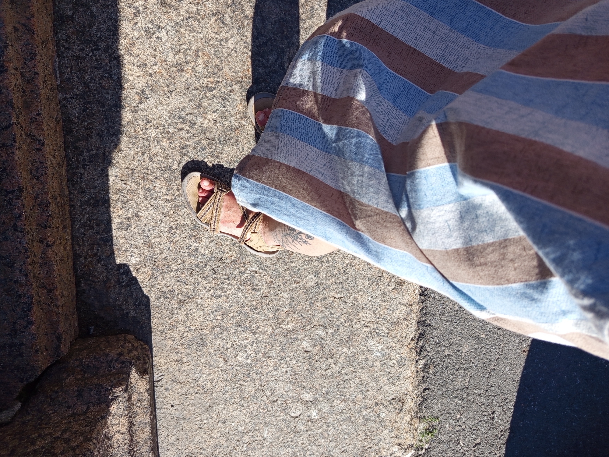 The image shows a person's legs and feet wearing sandals and a striped skirt, standing on a textured stone surface. The shadows suggest it is daytime. The area appears to be outdoors, possibly a park or historic site given the stone architecture visible in the background. There are no identifiable landmarks or features that would place this image specifically in Saint Petersburg, Russia.