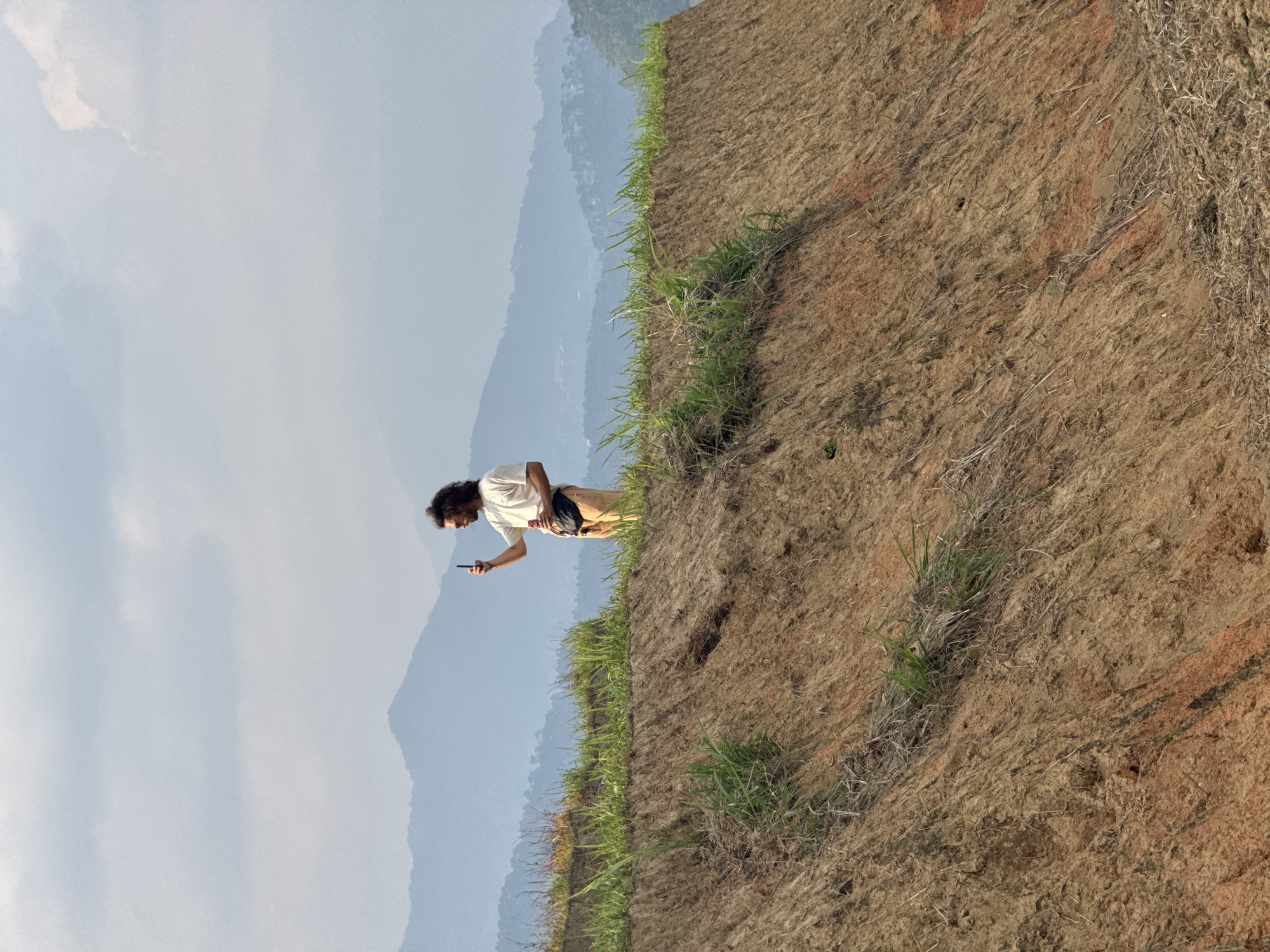 The image shows a person standing on a hillside, looking at a mobile phone. The hillside is covered in brown dirt and grass, and there are mountains in the background. The sky is partly cloudy. The location is in Pờ Ly Ngài Commune, Vietnam.