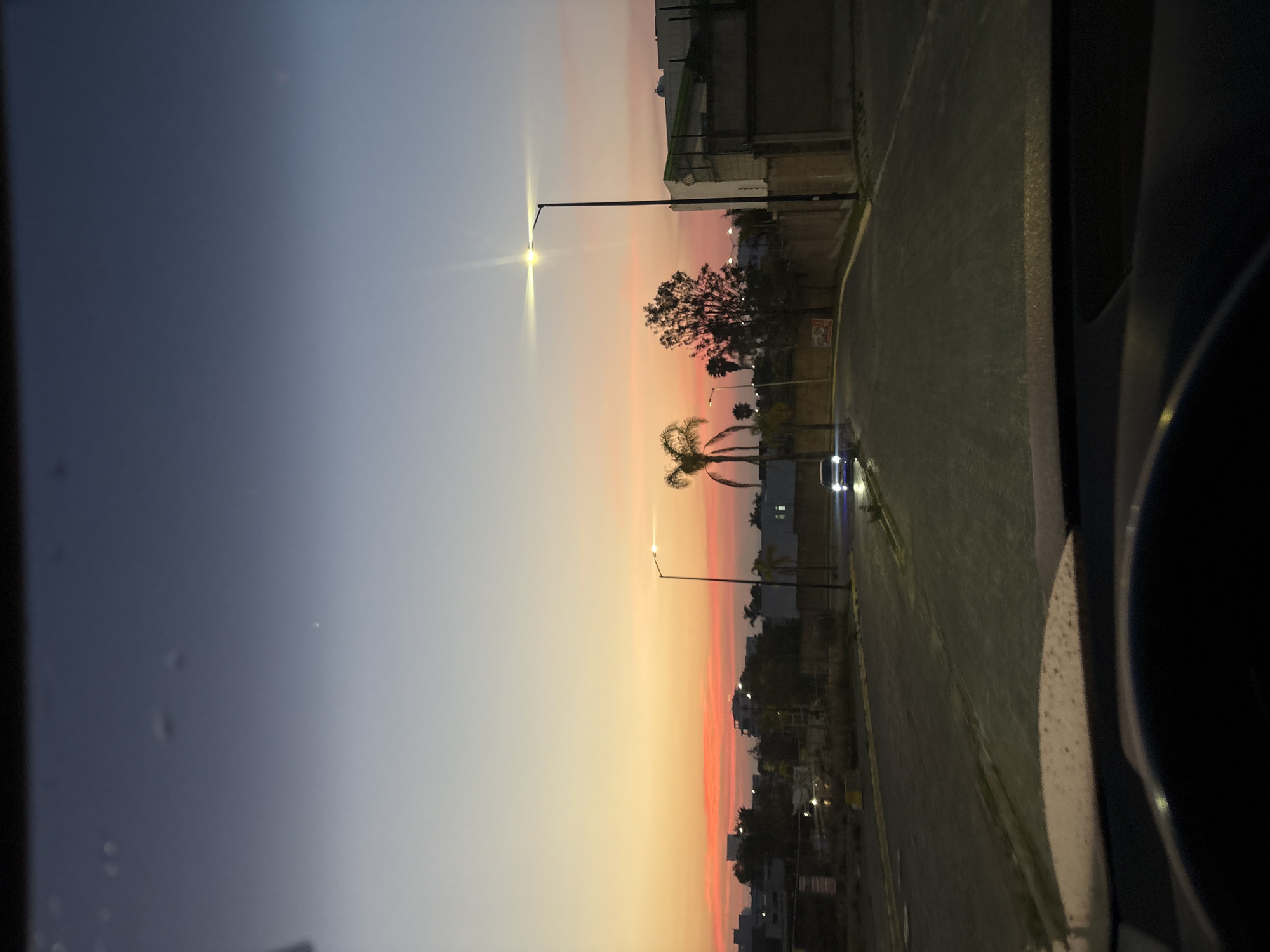 This image depicts a vibrant twilight scene over an urban or suburban street in San Bernardino Tlaxcalancingo, Mexico, viewed from inside a vehicle. The sky is a dominant feature, displaying a striking gradient from dark blue and purple at the top to warm shades of orange, pink, and yellow near the horizon, indicative of a sunset or sunrise.
A prominent tall street light with a bright white lamp stands in the midground, its light creating a noticeable lens flare. Several trees, including a large, leafy one and two distinct palm trees, are silhouetted against the colorful sky, lining what appears to be a paved road or street. Buildings with visible facades and dark outlines are present on both sides of the street. In the distance, the headlights or tail lights of another vehicle are visible on the road. A yellow line is discernible on the road surface, likely marking a lane or the curb. A concrete barrier or wall runs alongside a section of the street.
The foreground on the right side shows the dark interior frame of a vehicle, suggesting the photograph was taken from within a car. The vehicle's window appears to have small water droplets or smudges. The overall impression is one of a serene evening transition from day to night, with artificial lights beginning to illuminate the urban landscape as natural light fades. No legible text is visible in the scene.