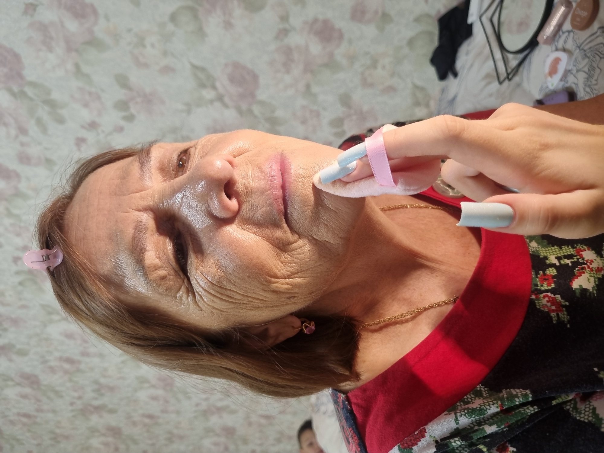A medium shot photograph shows an elderly woman applying makeup in a bedroom. The woman has short, light brown hair with a pink clip on top. Her skin shows wrinkles, and she has brown eyes. She is wearing a red and black patterned shirt with a red collar, and a gold chain necklace. Her left hand is holding a pink powder puff with a pink ribbon, which she is using to apply makeup to her chin. Her fingernails are painted a light blue color. In the background, a floral wallpaper is visible, and on a bed, there are some makeup items and a black fabric. A person is lying in bed in the background, out of focus. The lighting in the image is warm, with a strong highlight on the woman's face. The scene appears to be indoors. The image does not contain any visual indicators that would allow for its localization in the city of Chaikovsky, Russia.