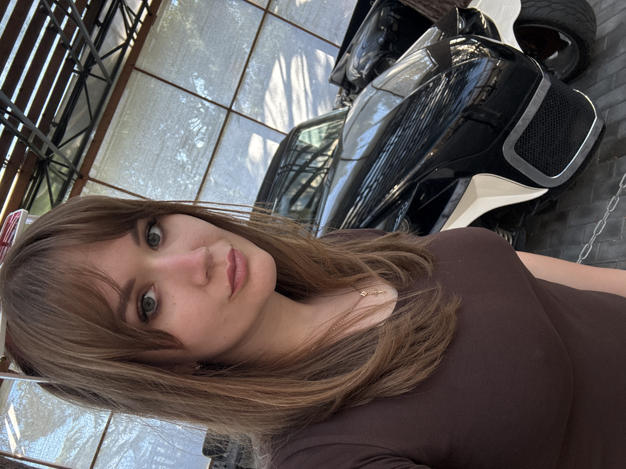 A young woman with shoulder-length brown hair and green eyes, wearing a brown long-sleeved top and a delicate gold necklace with a cross pendant, is positioned in the foreground. Behind her, partially visible, is a black and white three-wheeled vehicle. The background includes a building with a translucent roof structure featuring metal beams. Based on the prompt mentioning Ялта, Ukraine, the location is likely within this city.