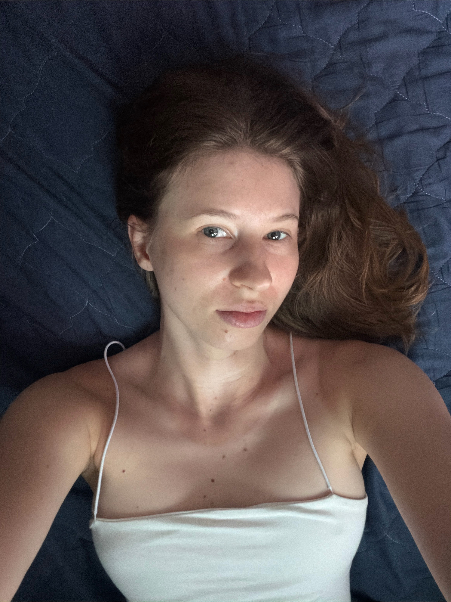 A woman with long, brown hair and fair skin is lying on a dark blue, quilted surface. She is wearing a white spaghetti strap top. Her head is tilted back and her eyes are looking towards the camera. The image appears to be a selfie taken from above. The background consists of the quilted surface, suggesting an indoor setting such as a bed or sofa. There is no visible indication of the city of Dongducheon-si, South Korea.