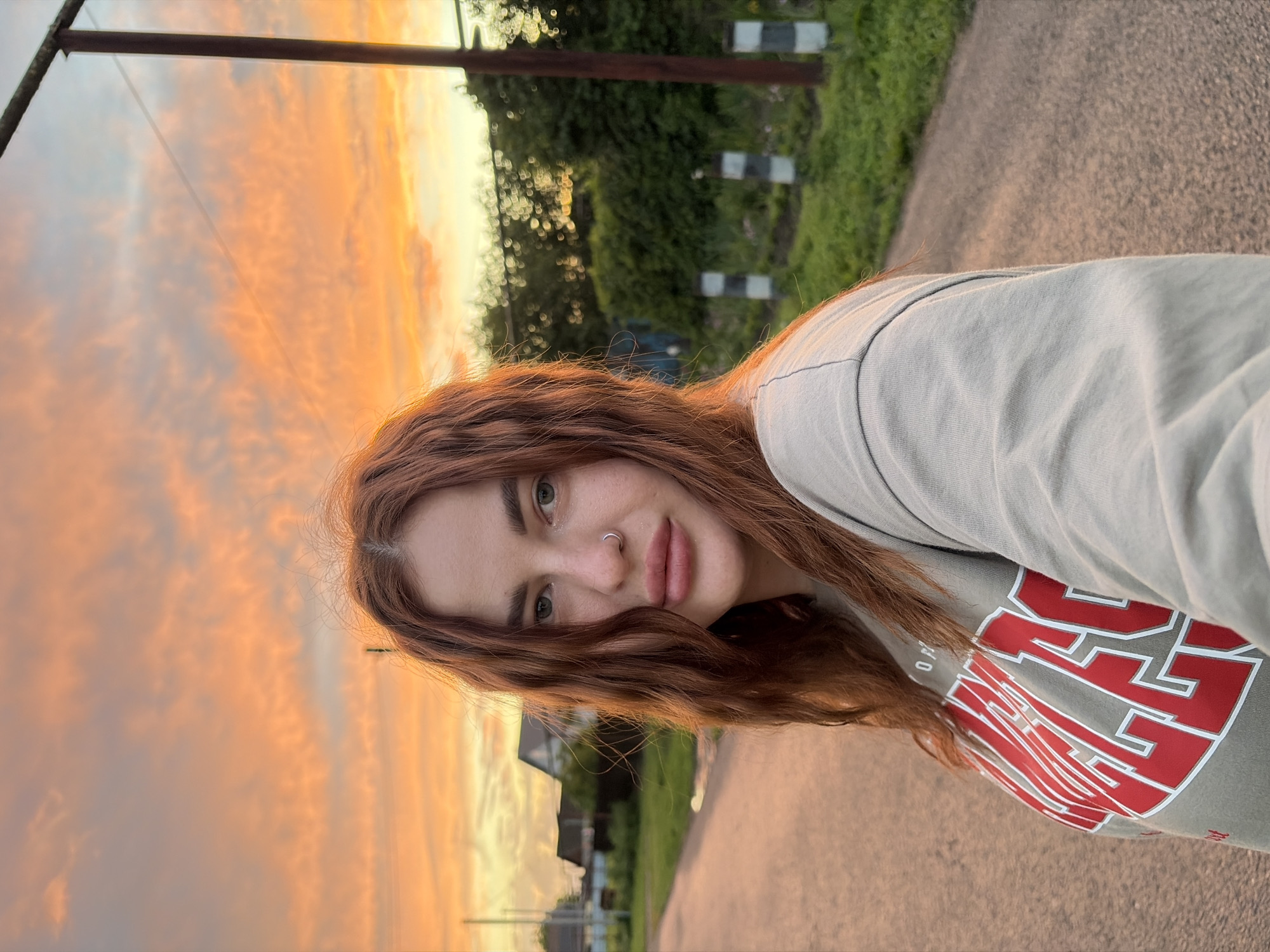 A woman with long, wavy, auburn hair is photographed outdoors. She wears a light gray, long-sleeved shirt with a large, red and white graphic on the front. Her skin tone is fair, and she has green eyes and a silver nose ring. The backdrop reveals a vibrant sunset with orange and yellow hues filling the sky. A utility pole stands tall on the left, and greenery with what appear to be striped poles lines the top of the frame. A paved road extends into the background, leading towards houses partially visible under the greenery. The location and city are not discernible from the image.