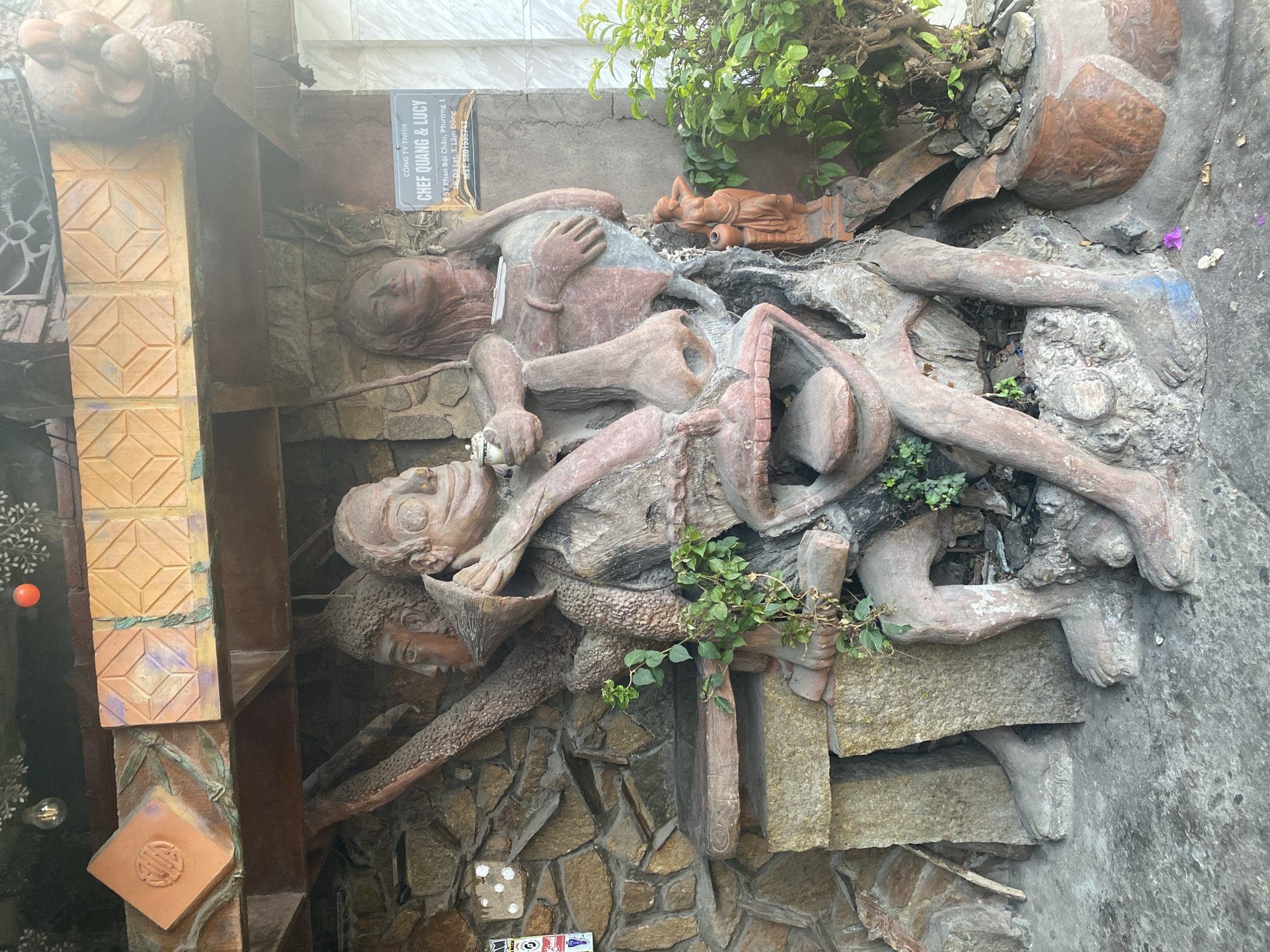 The image shows a concrete sculpture installation on the side of a building. The sculpture depicts multiple human figures in various poses, with one figure appearing to be reclining. The figures are crafted from a reddish-brown concrete-like material and are integrated into a textured background of stones and other materials. The installation also includes some plants and decorative elements. A sign is affixed to the wall nearby that reads "CHEF QUANG & LUCY" and includes an address: 57 Phan Boi Chau, Phuong 1, TP. Da Lat, T. Lam Dong.