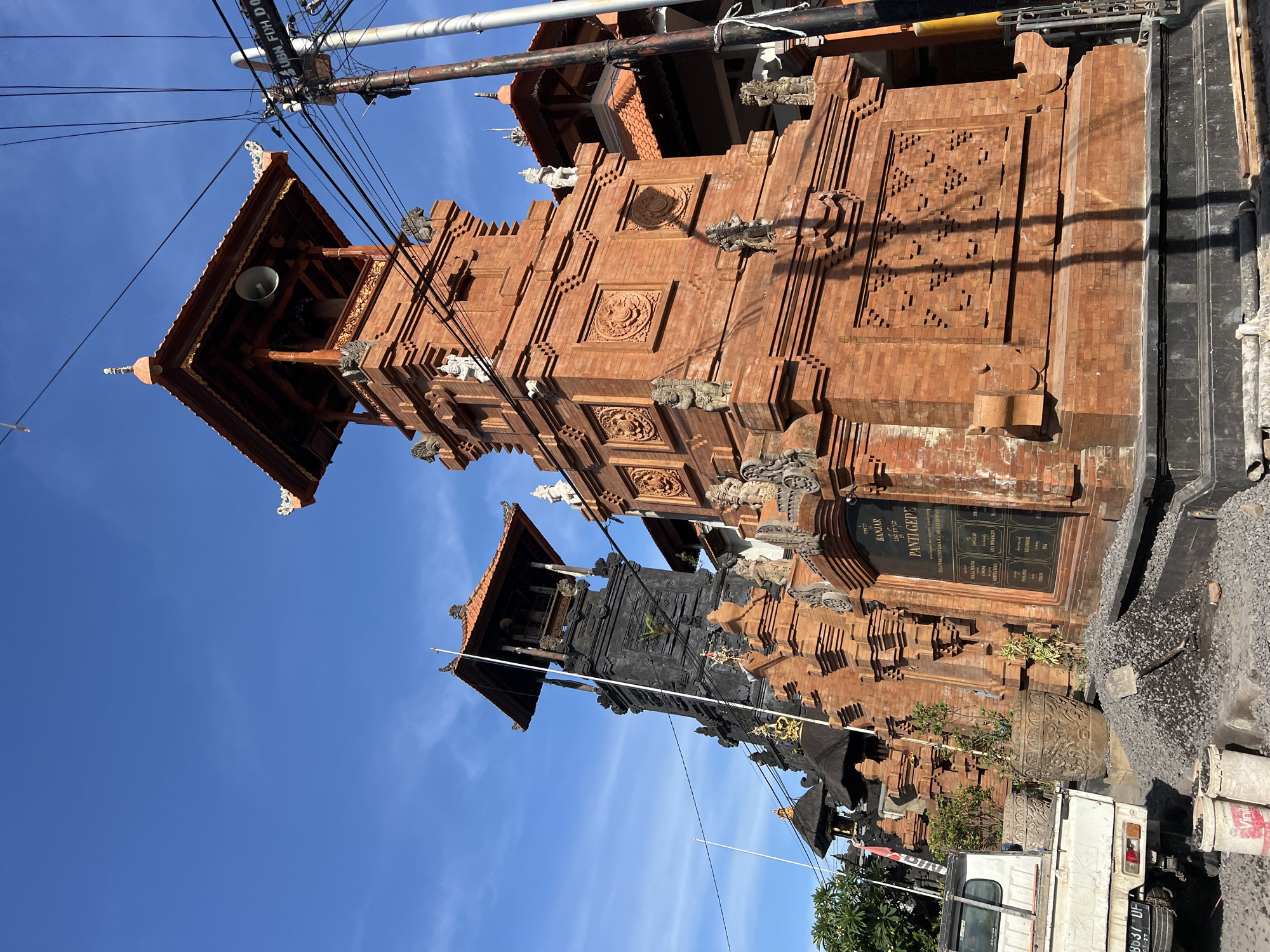 The image shows a Balinese Hindu gate structure, likely a Candi Bentar, under a bright blue sky with some wispy clouds. The gate is constructed of red bricks and adorned with intricate carvings and sculptures. Power lines and utility poles are visible above the structure. Part of the gate is painted black. A sign, reading "BANJAR PANDEGE", is affixed to the structure. A white truck is parked at the base of the structure. The gate is located within the city of Denpasar, Indonesia.