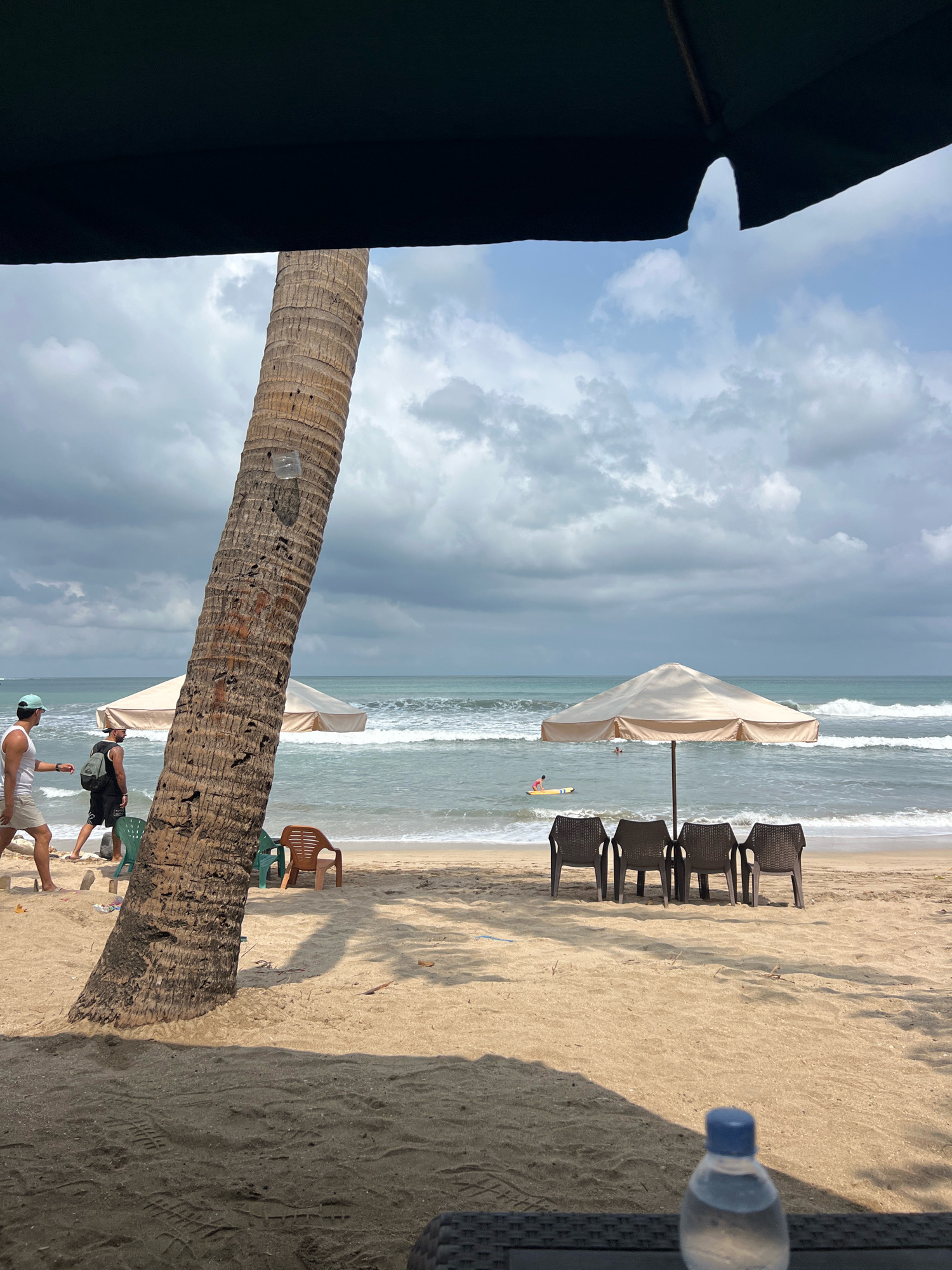 This image captures a view of Kuta Beach in Bali, Indonesia, from beneath a large dark umbrella, possibly from a beachfront cafe or restaurant. The foreground is dominated by the textured sand and a dark woven surface, likely a table, with a partially visible plastic water bottle with a blue cap in the lower right corner.
A prominent, textured palm tree trunk leans diagonally from the left mid-ground towards the foreground, casting a shadow on the sand. To the right of the palm tree, a beige beach umbrella stands over a row of four dark plastic chairs arranged on the sand.
In the mid-ground, the ocean stretches to the horizon under a cloudy sky. Waves are breaking on the shore, indicating active surf. A person on a yellow surfboard is visible in the water near the umbrella. Two individuals are walking on the beach in the left mid-ground, one wearing a white tank top and khaki shorts, the other a black tank top and shorts with a backpack. Scattered green and brown plastic chairs are also visible on the sand. Another beige umbrella is positioned further back on the beach, to the left of the main umbrella. The scene suggests a slightly overcast day, common in Kuta, with typical beach amenities and activity.