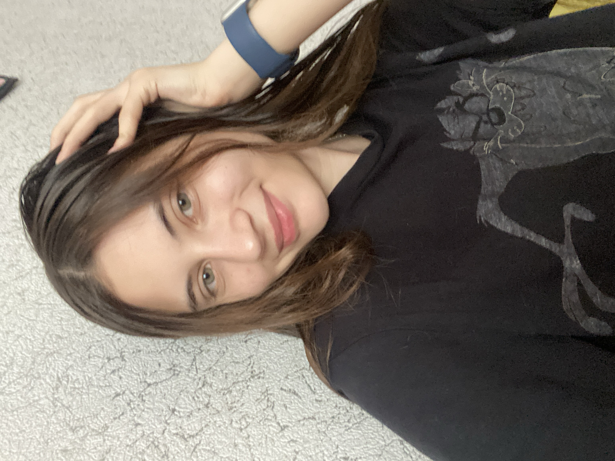 A young woman with long, dark brown hair and green eyes is captured in a selfie. She is wearing a black t-shirt with a graphic print of the Tazmanian Devil. A blue smartwatch is visible on her left wrist. The background appears to be a textured gray wall. The image does not contain any information that would indicate its location in Votkinsk, Russia.