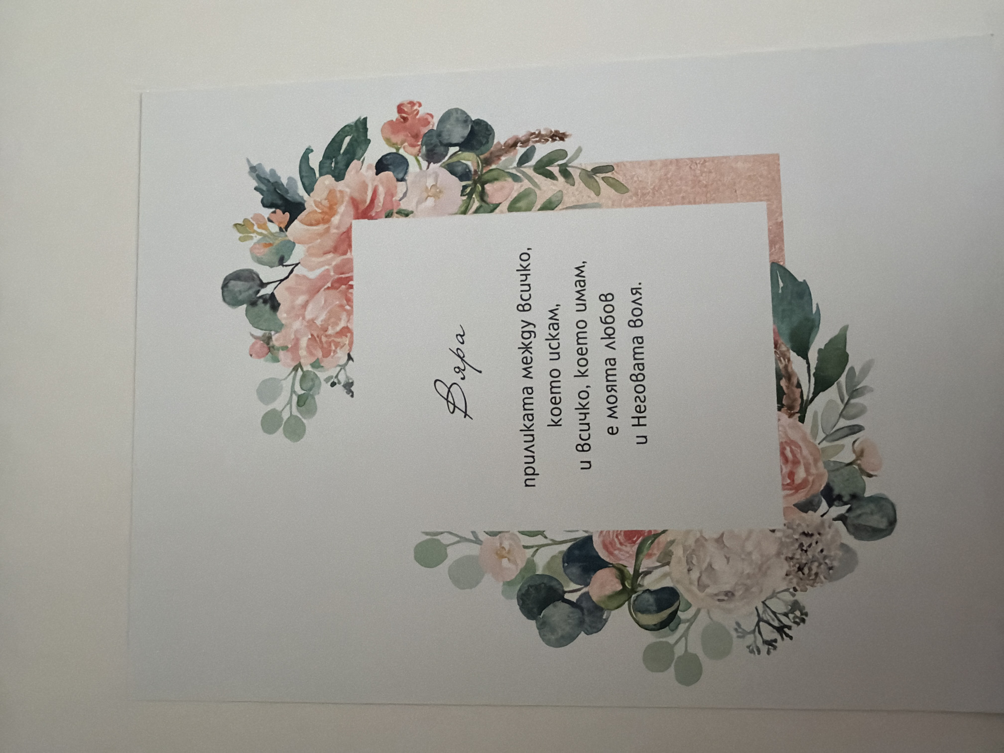 The image is a close-up, slightly angled shot of a greeting card. The card features a floral design with watercolor illustrations of pink roses, eucalyptus leaves, and other greenery arranged in a bouquet-like fashion around the edges. In the center of the card, there is a white rectangular area with text printed in black. The text is in Bulgarian and appears to be a quote or message. The word "Вяра" (which translates to "Faith" in English) is written in a script font above the main body of text. The background of the card is white, and the overall presentation is delicate and artistic. The image is taken indoors, likely in a home or studio setting, as indicated by the plain, neutral background which appears to be a wall or surface. There is no information available to determine the location in the city of Pazardzhik, Bulgaria.