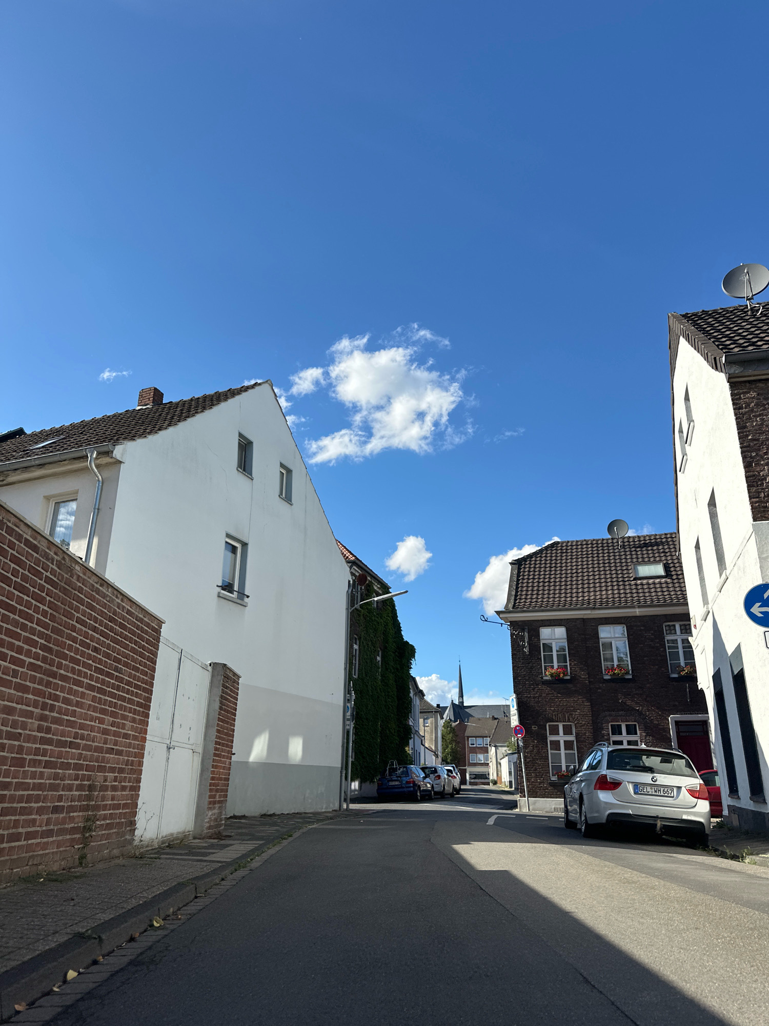 The image shows a street in Krefeld, Germany, on a sunny day with a clear blue sky and a few white clouds. The street is lined with residential buildings and parked cars. On the left side of the street, there is a white house with a red brick wall in front of it. The house has several windows and a gable roof. Further down the street on the left, there is a building covered in ivy. On the right side of the street, there is a dark brick building with white trim around the windows. A silver BMW station wagon is parked in front of this building, its license plate is visible. Further down the street, other cars are parked and buildings can be seen. A street sign with a left arrow is visible on the right side. The road surface is asphalt, with a visible curb and sidewalk.