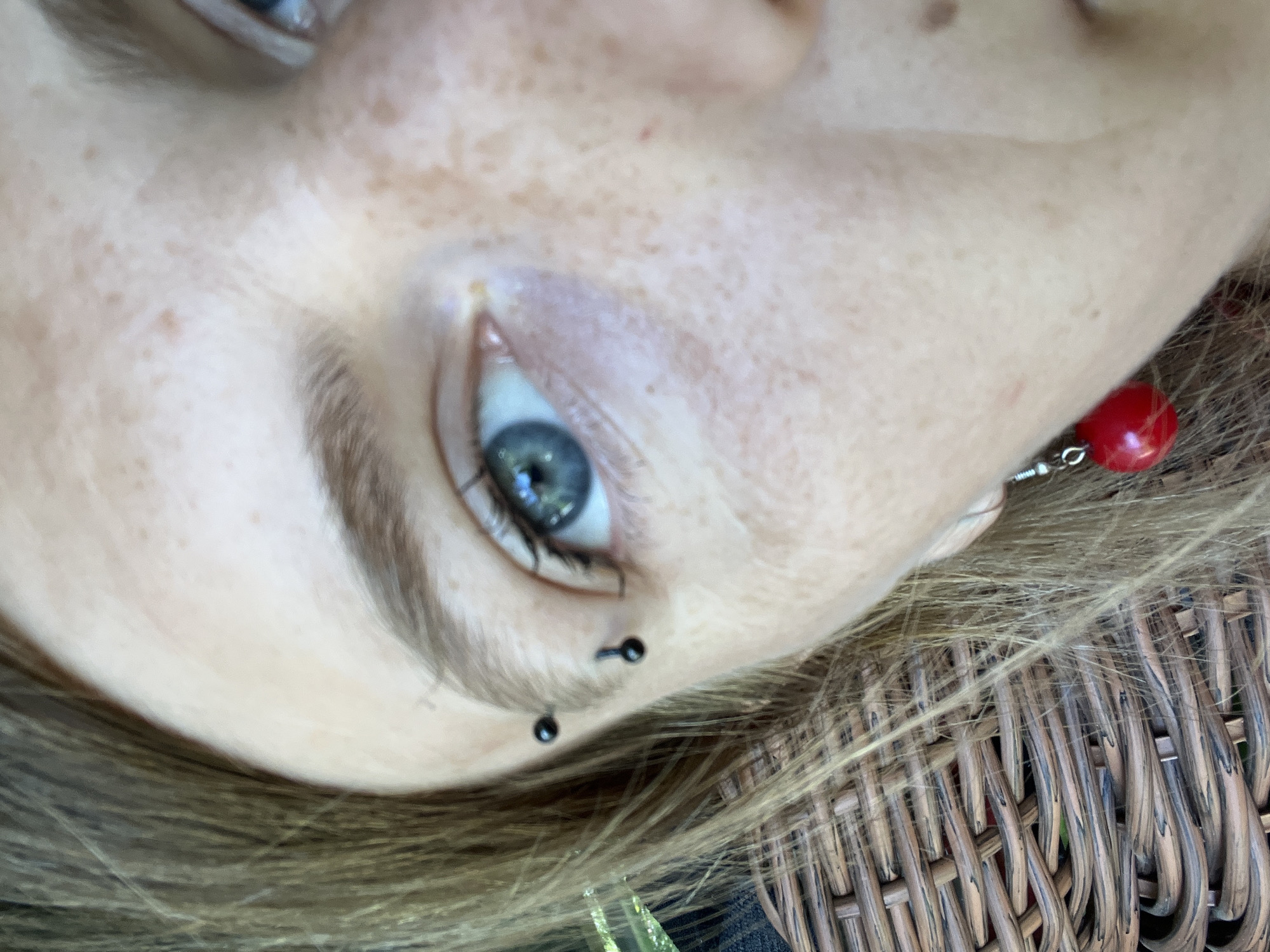 The image is a close-up, eye-level shot of the right side of a person's face, focusing on their eye and eyebrow area. The person has light-colored skin with freckles. Their right eye is blue and appears to be looking slightly off-camera. The eyebrow is light brown and arched. There are two black hoop piercings in the eyebrow and one in the upper cheekbone below the eyebrow. The person is wearing a red, spherical earring that dangles from their earlobe. Strands of light brown hair are visible on the left side of the frame, some of which are resting on a woven wicker surface. The overall lighting suggests an outdoor setting with natural light. The image was taken in Дунілавіцкі сельскі Савет, Belarus.