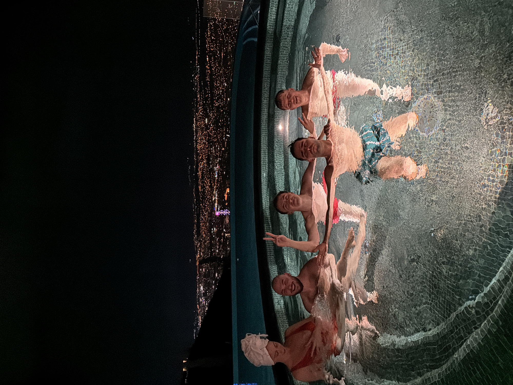 Five people are in a pool at night overlooking a city at night. The pool is surrounded by tile. The people are smiling and making hand gestures. The city is in Tbilisi, Georgia.