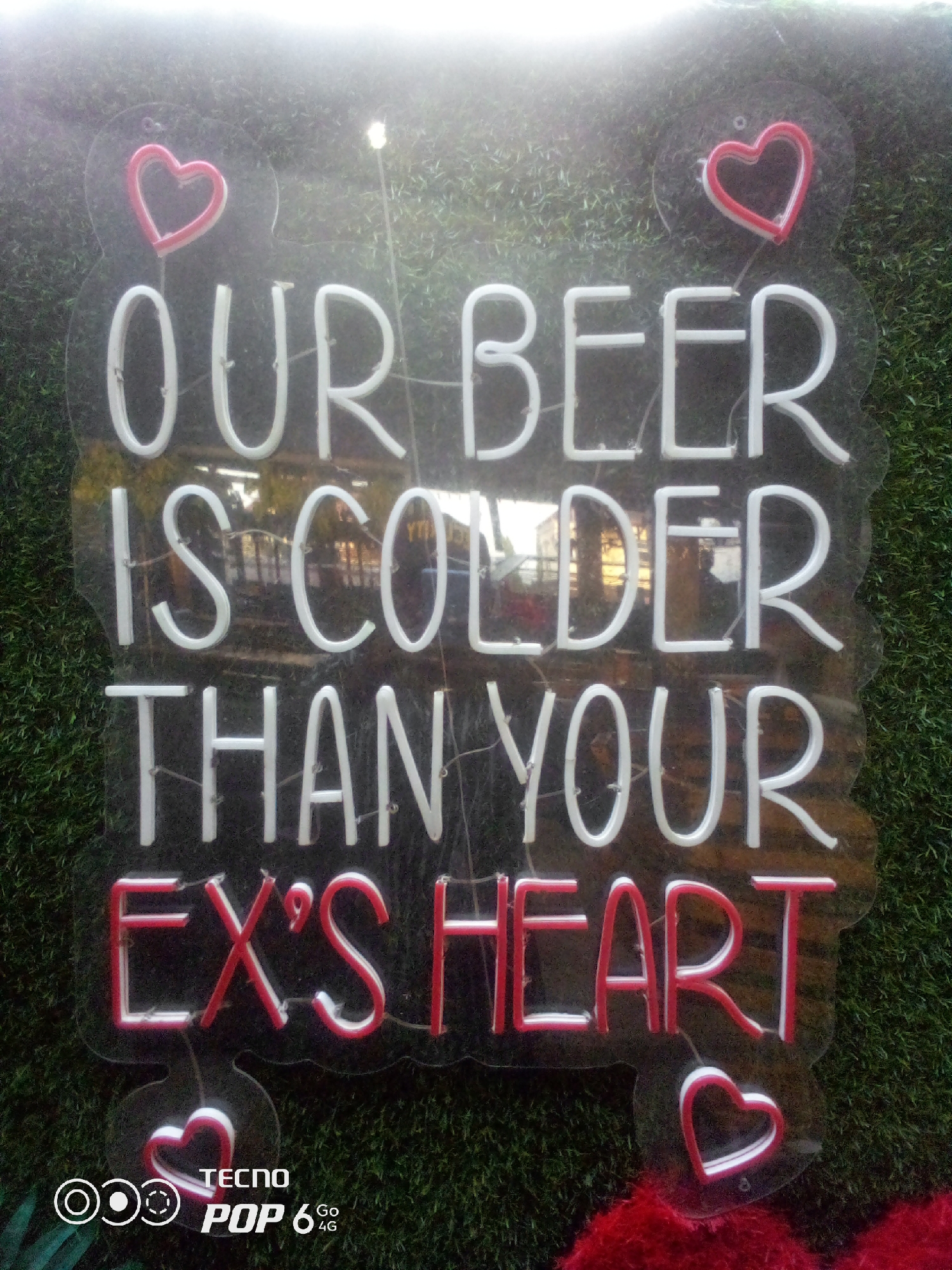 In Howayi, Nigeria, a neon sign is displayed against a backdrop of artificial turf. The sign features the text "OUR BEER IS COLDER THAN YOUR EX'S HEART" in white neon letters. Two red and white neon hearts are positioned at the top left and bottom right corners of the sign. The sign itself is mounted on a clear acrylic backing. A small portion of a red, fuzzy material is visible at the bottom right of the image, and a watermark for "TECNO POP 6 4G" is present in the bottom left corner. The lighting in the image is bright, creating a halo effect around the neon elements and obscuring some of the background details.