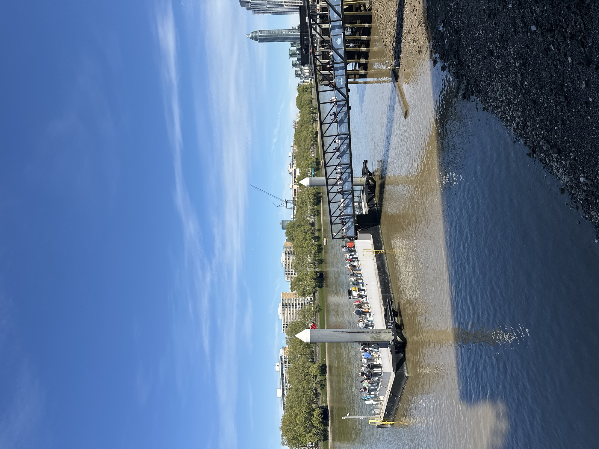 The image shows a view of the River Thames in London, United Kingdom. A metal pier extends from the riverbank, with a walkway leading to a docking area. Several boats are moored at the pier, some containing people. A black barrier is visible at the water’s edge. Buildings and trees line the riverbank in the distance, and a construction crane is visible. The sky is clear and blue. The image is rotated counterclockwise.