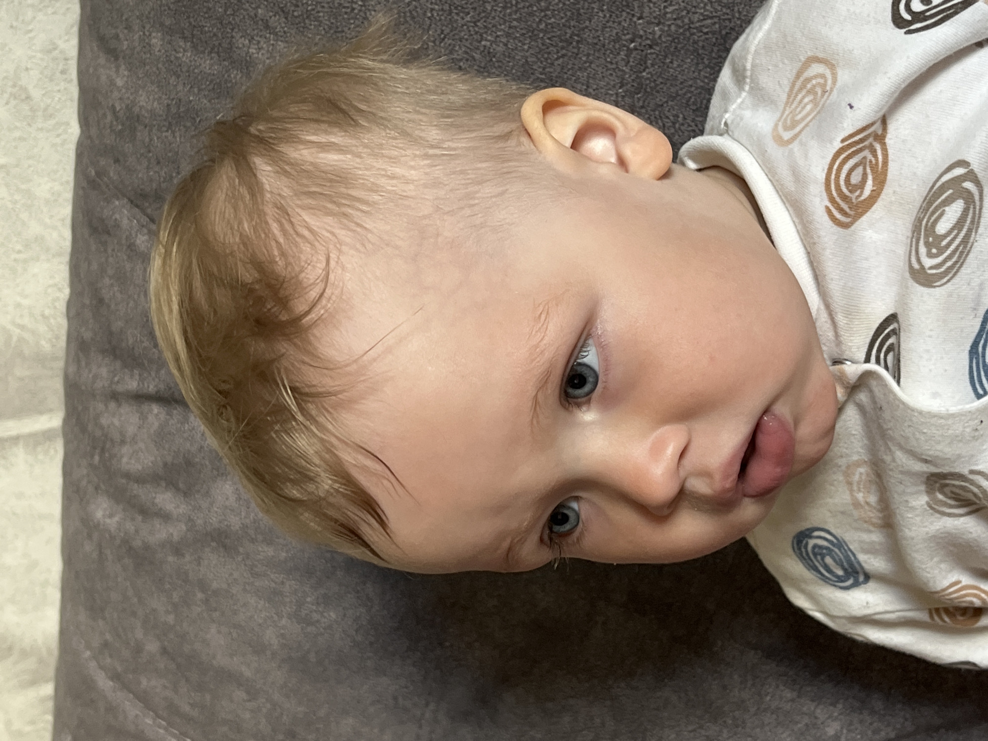 A young child is lying on a grey pillow, wearing a white shirt with circular patterns in shades of brown and blue. The child has fair skin, blue eyes, and light brown hair. The child's mouth is slightly open, with the tongue visible. The image does not provide information on the child's location in Chaikovsky, Russia.