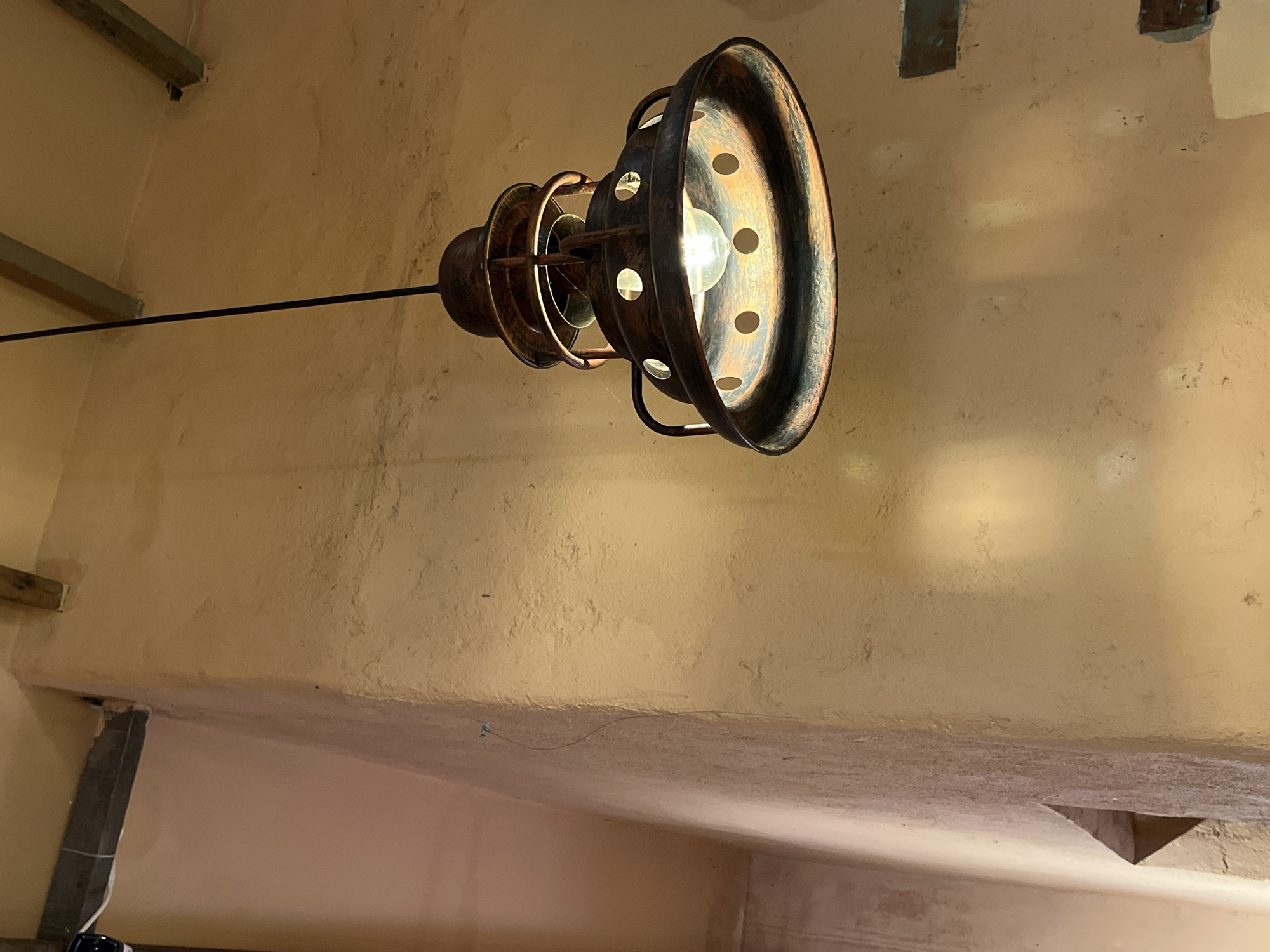 A low-angle shot shows a hanging, industrial-style light fixture attached to a black metal rod against a textured, beige wall. The fixture is composed of a series of concentric metal rings with holes, revealing a lit bulb in the center. Patches of discoloration and markings are visible on the wall's surface. The lower portion of the image shows a pinkish-beige ceiling with beams and posts. No people are present. The location is not evident from the image.