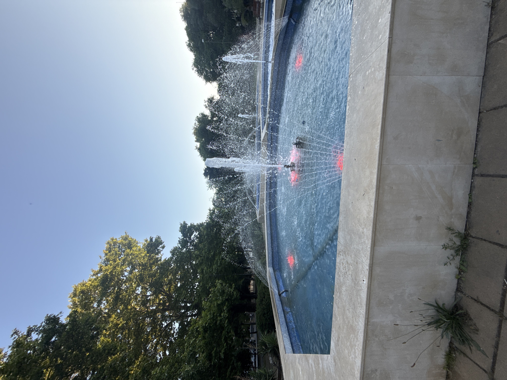 A fountain with multiple jets of water spraying upwards is the main subject of this image. The fountain is situated in a pool with blue tiled edges. Underwater red lights are visible in the pool, casting a glow on the water. The fountain is surrounded by a light-colored stone border, and beyond that, paved ground. Lush green trees are visible in the background, suggesting an outdoor setting, likely a park or public space. The sky is clear and blue, indicating it is daytime. The sun is shining, casting bright light on the scene. The image appears to be taken from a slightly elevated angle, looking down into the fountain. There is no visible text. The overall impression is one of a pleasant, sunny day in a well-maintained outdoor area.