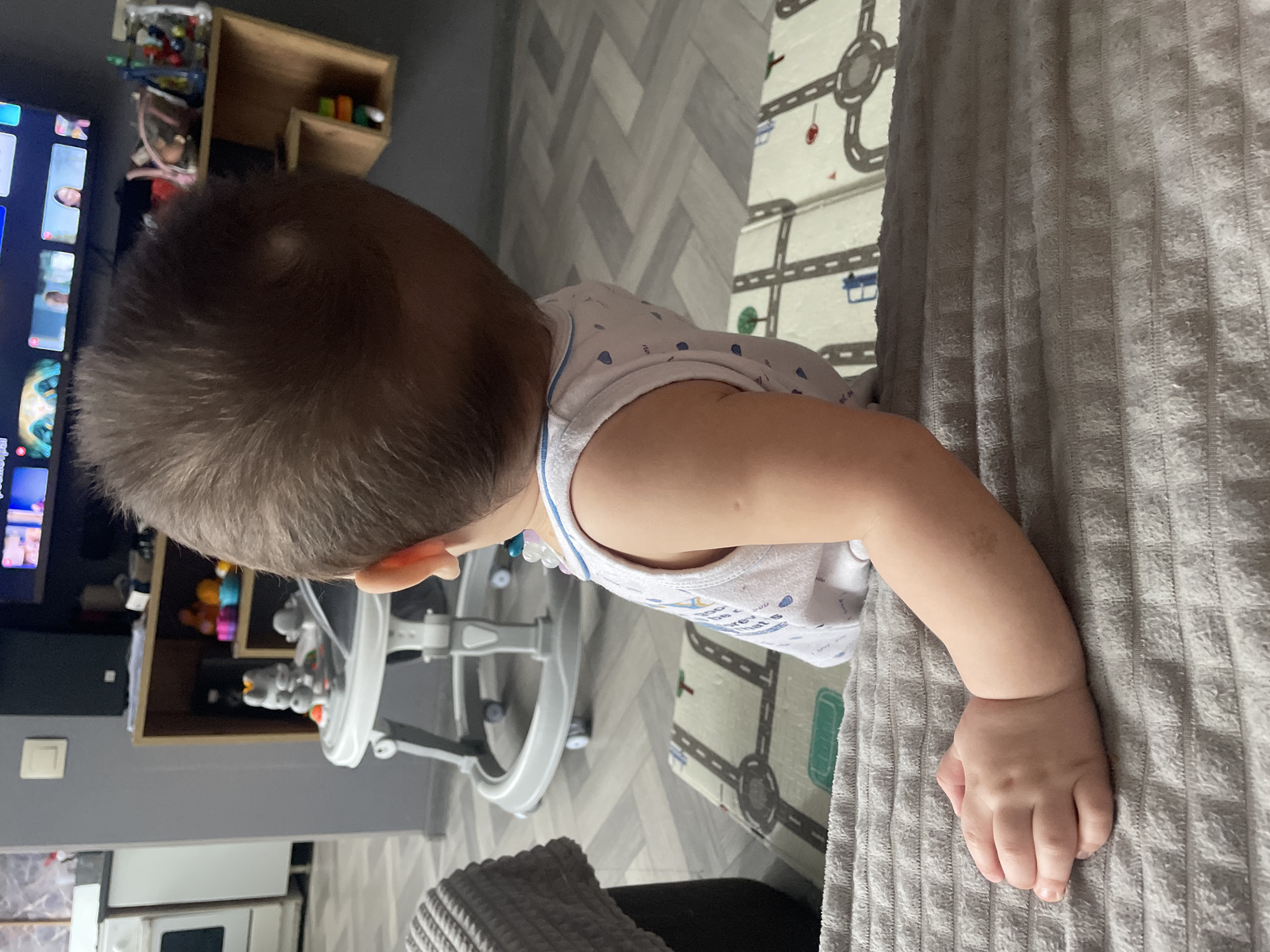 A baby wearing a white and blue top is lying on a gray textured surface. In the background, there is a play mat with a road design, a white walker, a TV and shelf with toys and an appliance (refrigerator).