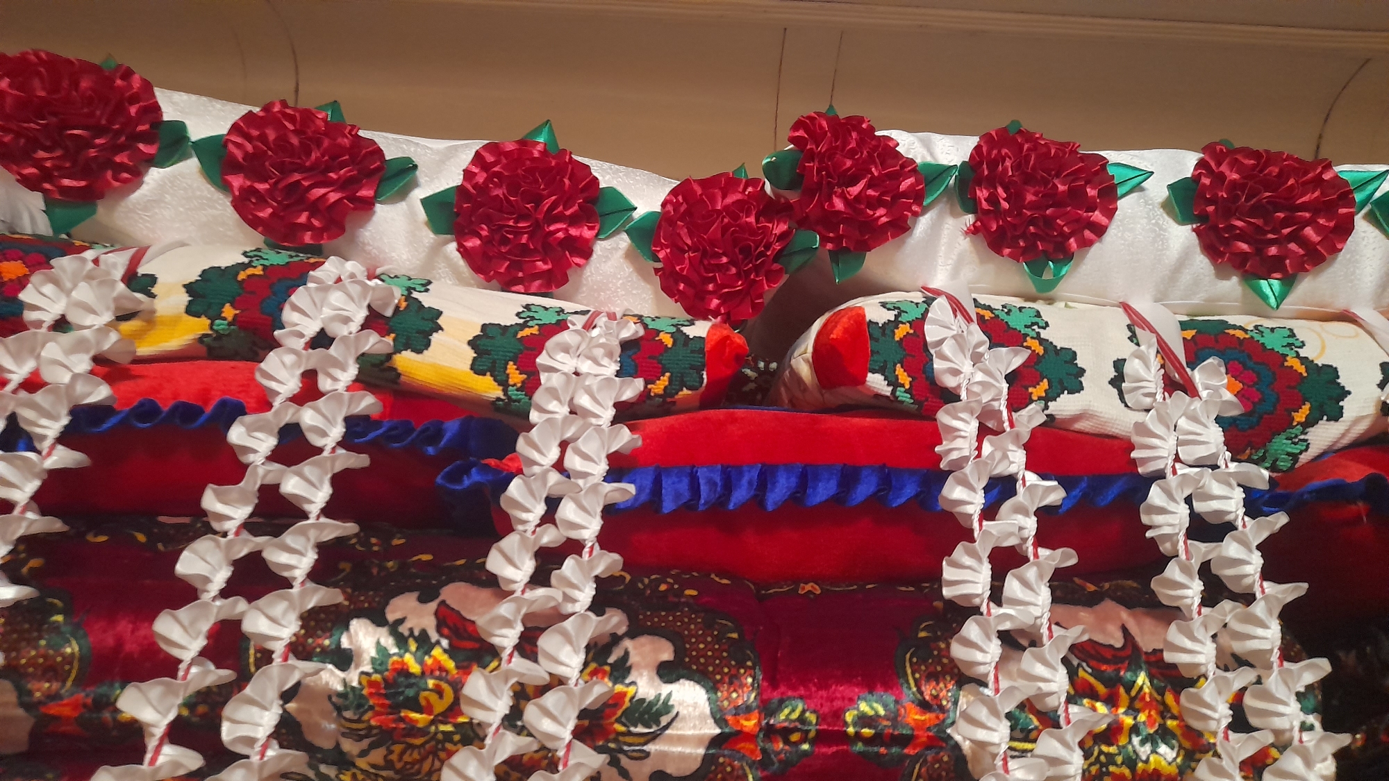 The image shows a close-up view of stacked decorative pillows and bolsters. The top layer consists of white pillows adorned with red fabric flowers with green ribbon leaves. Below these are several cylindrical bolsters covered in patterned fabric featuring intricate designs of red, green, yellow, and blue motifs. Between the bolsters, there are red and blue ruffled fabric edges. White ribbon garland-like decorations are draped in front of the lower layers of pillows and bolsters. The overall impression is one of festive or ceremonial decoration. The image does not contain any discernible features that would indicate its location within the city of Osh, Kyrgyzstan, or any specific environmental context beyond the immediate arrangement of decorative items. There are no people or actions depicted.