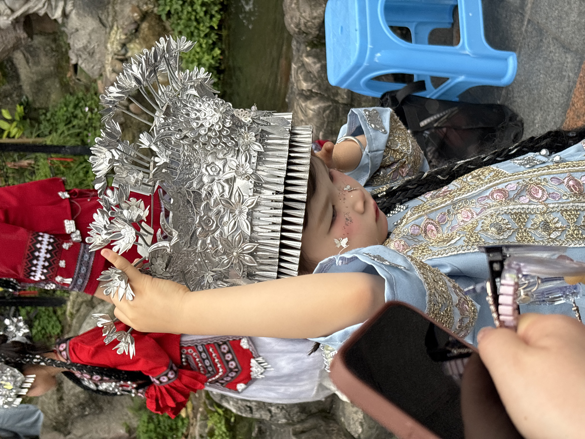 A young person in traditional silver and red clothing and a large silver headpiece poses for a photograph. A hand holding a phone is partially visible in the foreground. A blue plastic stool, a black bag, and parts of a body of water are visible in the background. The photograph appears to be taken outdoors in a garden or park-like setting. This event occurred in Yuzhong District, China.