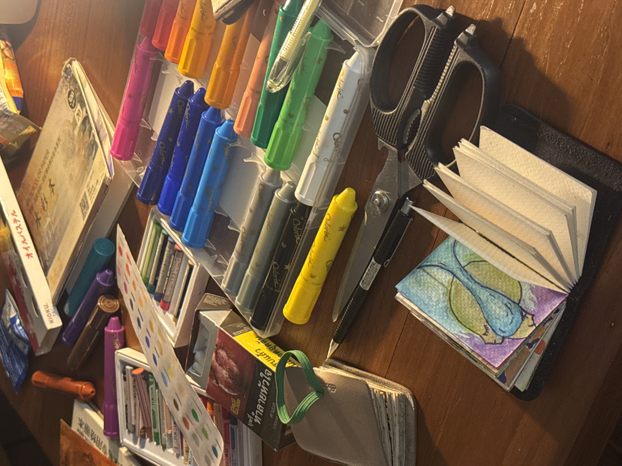 This image captures a close-up, overhead view of an art supply assortment laid out on a wooden surface, likely a desk. The scene is lit by warm, directional light, casting distinct shadows and suggesting it might be late afternoon or evening.
The subjects are primarily art supplies, including a variety of markers, crayons, and pastels in a spectrum of colors. Several items are neatly organized in clear plastic containers, while others are loose. Notable objects include a pair of black scissors, a mechanical pencil, and several small sketchbooks or notebooks. One sketchbook is open, displaying colorful, abstract watercolor drawings. Another features a textured cover with a green elastic loop. A box with Thai text, potentially cigarettes, is also visible among the art supplies.
The overall impression is one of creative potential and artistic endeavor. The scattered nature of some items suggests they are in active use or have been recently used. The wooden surface provides a warm, rustic backdrop for the vibrant art materials. The visible text includes brand names like "KIOKU" and "Colorix," as well as Japanese and Thai script, with one prominent Thai phrase translating to "Camel cigarettes." The setting appears to be indoors, given the controlled lighting and the presence of art materials, and the mention of "Chiang Mai City Municipality, Thailand" in the prompt suggests a local context for these items. There are no people or animals visible in the image.
