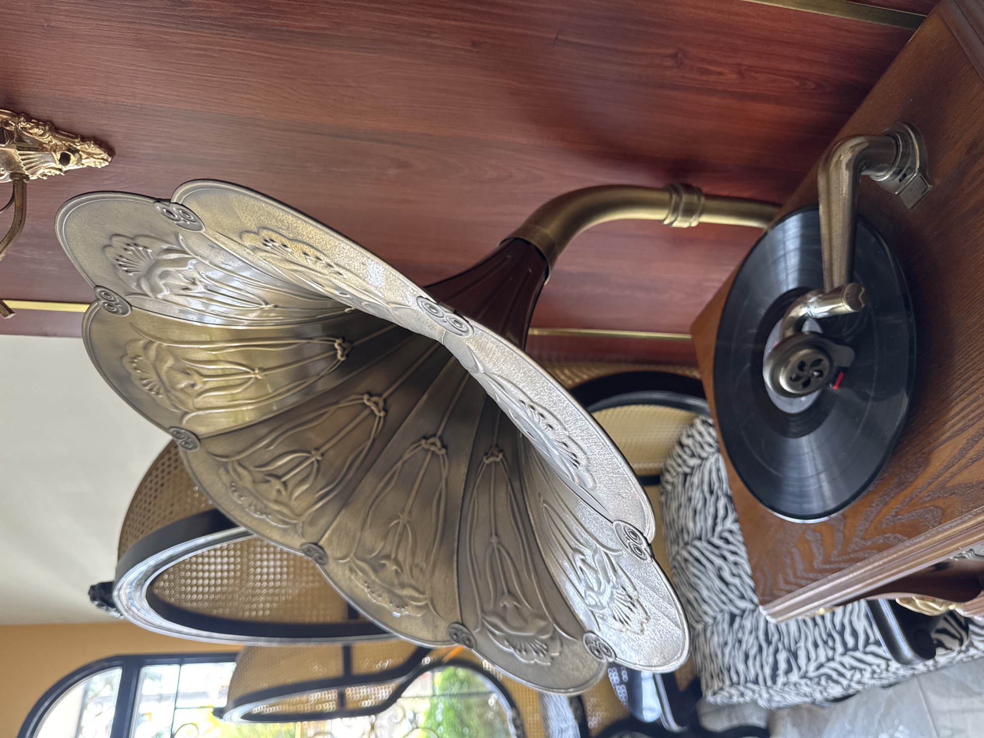 The image depicts a vintage gramophone with a large, ornate horn. The horn is metallic with a decorative floral pattern. The arm connecting the horn to the turntable is brass. A black vinyl record sits on the wooden turntable. The gramophone is positioned near a wicker chair and a zebra-patterned cushion. The location is Đà Nẵng, Vietnam.
