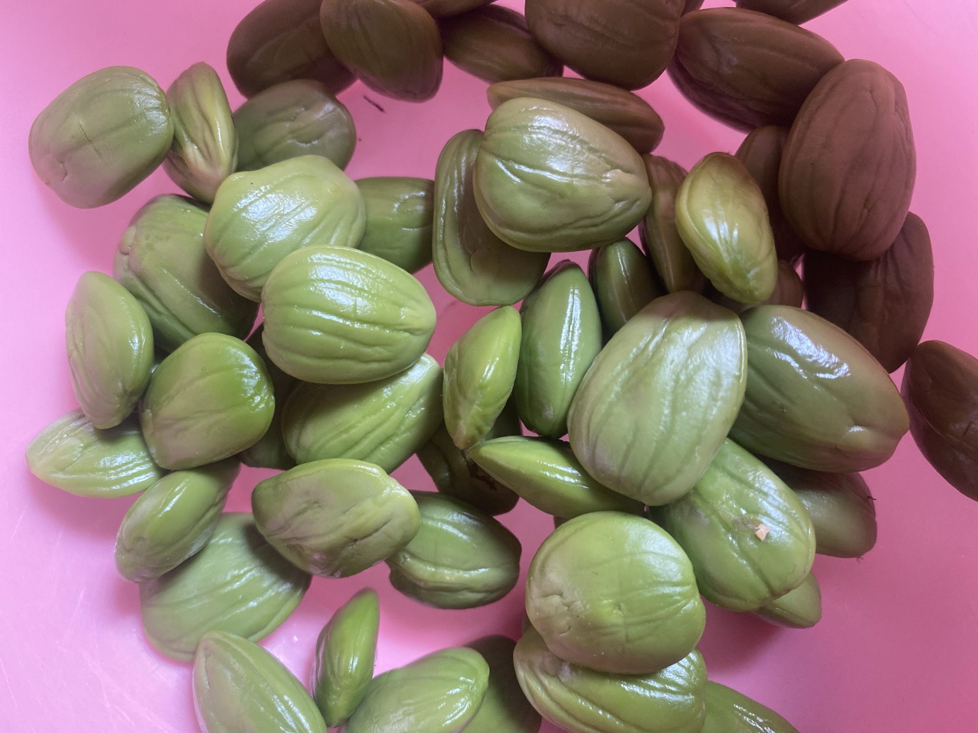 A close-up, overhead shot shows a pile of bright green, oval-shaped legumes, likely Parkia speciosa, commonly known as petai or stink beans, in a pink bowl. The beans are smooth and plump, with visible ridging on their surface. Some beans are stacked on top of others, creating a sense of abundance. The background is a plain, vibrant pink, which contrasts sharply with the green of the beans. The lighting is bright, casting subtle shadows and highlighting the glossy texture of the legumes. The image is sharp and detailed, capturing the natural form and color of the beans. The location is described as Malinau Kota, Indonesia.