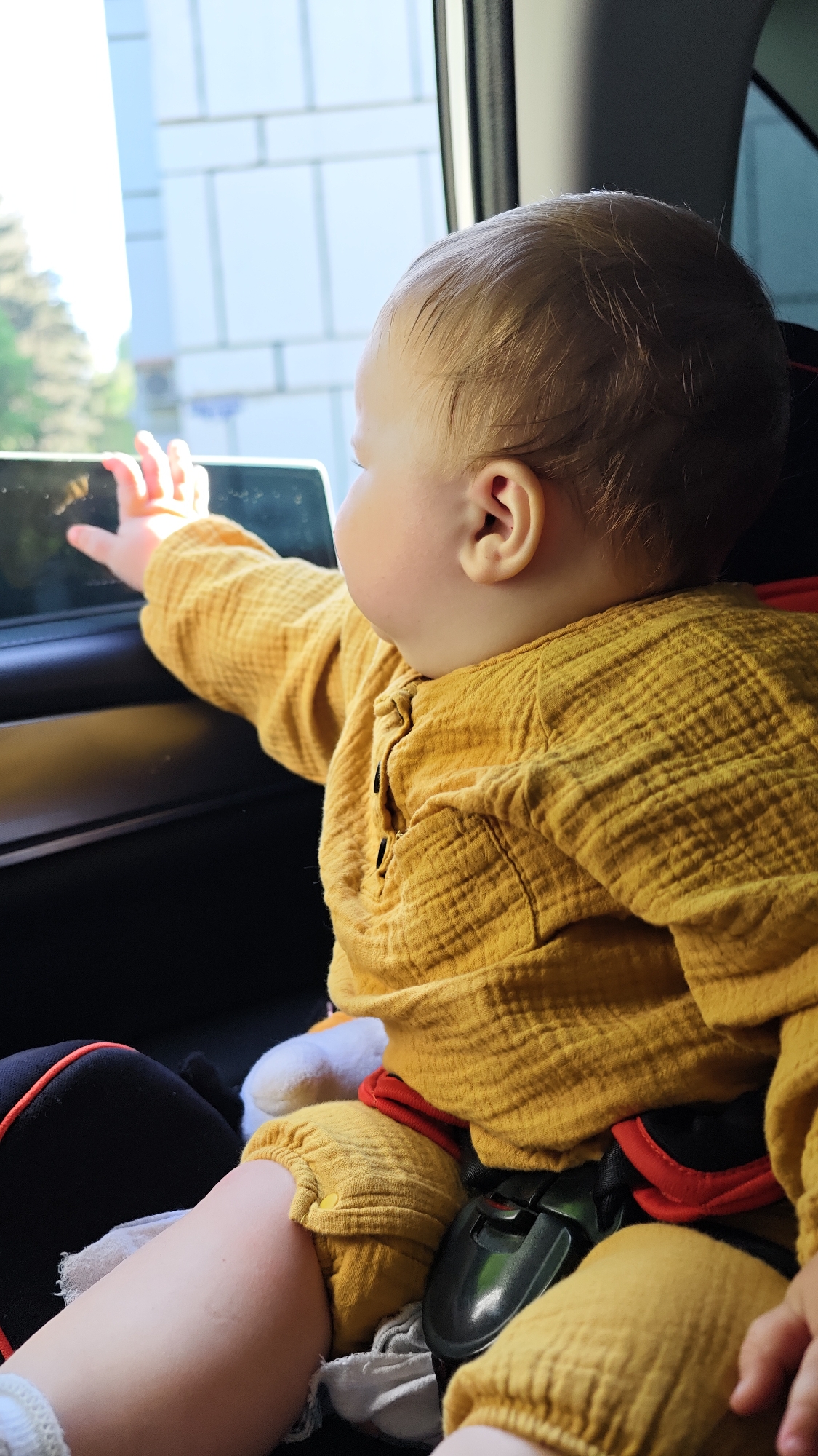 A baby is seated in a car seat, secured with a safety belt. The baby is wearing a yellow shirt and pants. The baby's left arm is extended towards the car window. Outside the window, the trees and building facade are visible, indicating the car is in motion. The car appears to be moving in the city of Chaikovsky, Russia.