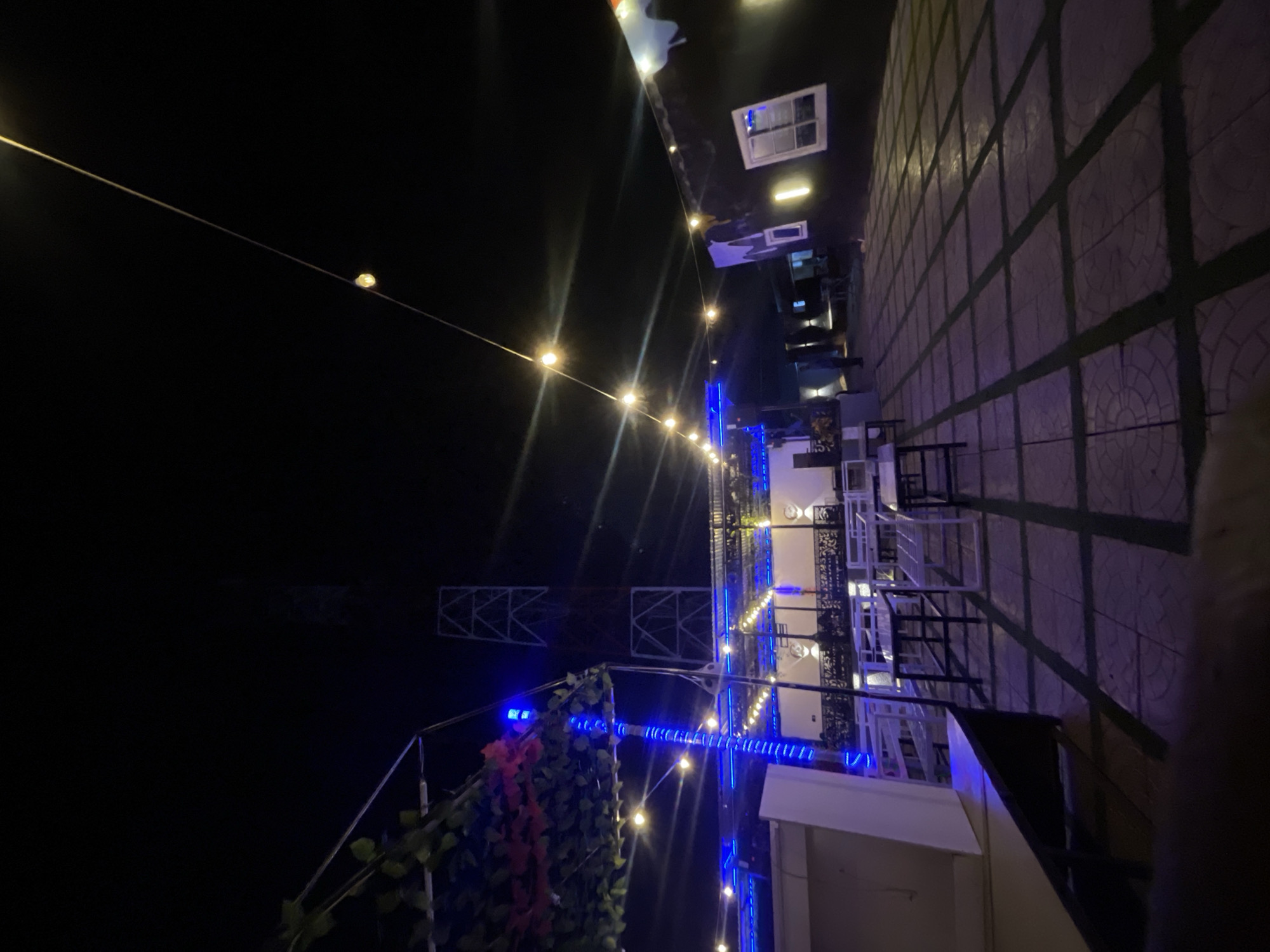 The image depicts a nighttime scene at an outdoor or rooftop bar or restaurant, identified as Bar Jirgi Summa in Nigeria. The setting is characterized by its dynamic artificial lighting, creating a vibrant atmosphere.
Overhead, warm yellow string lights are strung across a dark, open space, with a distinct light bulb visible on a diagonal wire in the upper left. These are complemented by cool blue LED strip lights that are wrapped around structural elements, possibly railings or beams, providing a modern contrast. More string lights are visible further into the establishment.
The main area features numerous tables and chairs, some white and some dark, arranged on a patterned tiled surface that appears to be a patio or floor. This surface is delineated by a grid of darker lines. In the foreground, green foliage, possibly vines, with accents of red, drapes over a support structure, adding a natural element. In the background, dark, skeletal structures with square metal frames are visible against the night sky, suggesting part of the building's architecture or a decorative framework. Small, illuminated window-like elements and reflections are also present on a dark vertical surface.
Although no people are visible, the arrangement of seating and the lighting suggest a venue designed for social gatherings and evening entertainment. The overall mood is one of an inviting, illuminated space ready for patrons. There is no discernible text in the image.