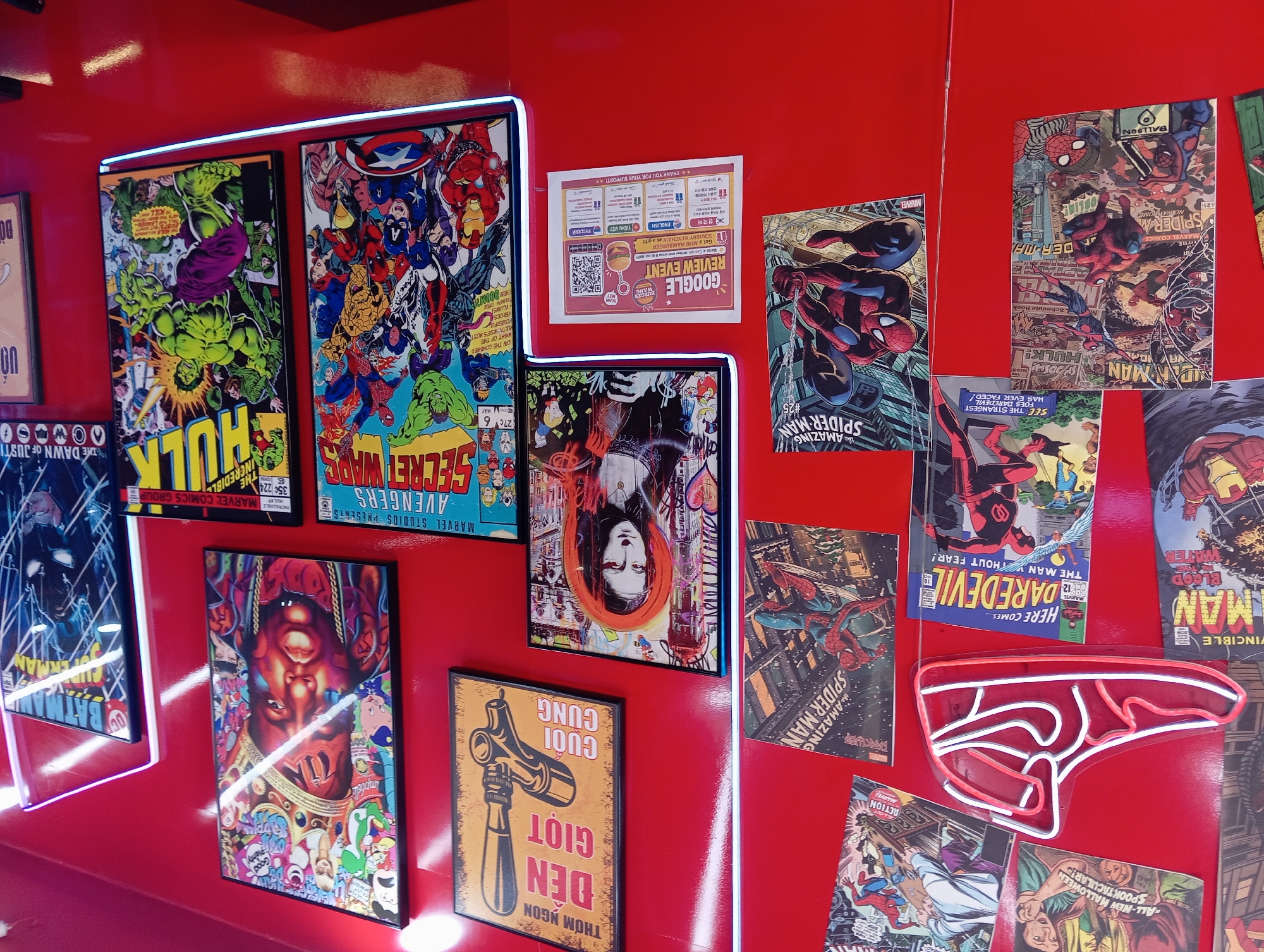 The image depicts a brightly decorated wall, likely in a restaurant or cafe, with numerous framed comic book covers and posters adorning a vibrant red surface. Dominating the left side is a neon sign shaped like a red Nike sneaker. Various Marvel comic book covers, featuring characters like Spider-Man and Daredevil, are prominently displayed. There are also posters of famous figures, including a stylized portrait of Biggie Smalls and a Mona Lisa artwork with a graffiti-like style. Another framed piece shows a beer tap with Vietnamese text. In the lower section, a Google review event flyer is visible. The lighting is consistent with an indoor setting, with some areas highlighted by neon light strips along the bottom edge of some frames. The overall impression is one of a pop culture-themed establishment. The specific location is a Burger Wang restaurant on To Hien Thanh Street in Nha Trang, Vietnam.