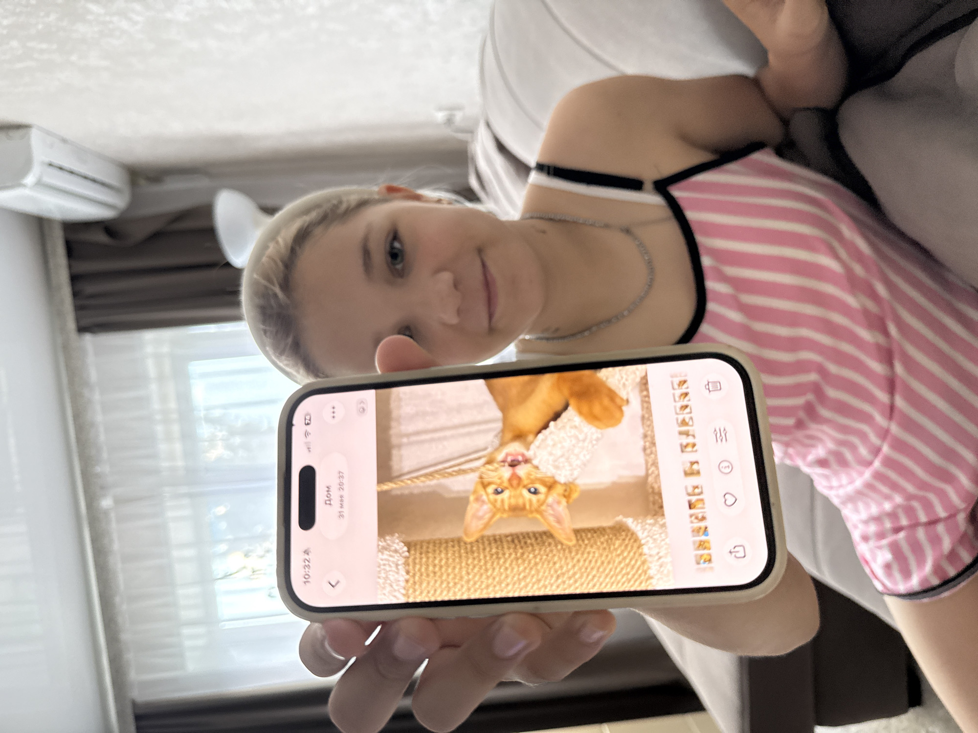 A young blonde woman wearing a pink and white striped camisole and a silver necklace is sitting on a sofa, holding up a smartphone. The smartphone displays a photo of a ginger cat with its mouth open, perched on a scratching post. The woman is looking at the camera and smiling. The background shows a living room with curtains and an air conditioner. The timestamp on the phone indicates it is 10:32 AM and the date is May 31st, 20:37, which is likely a typo for 8:37 PM. The location is not specified in the image.
