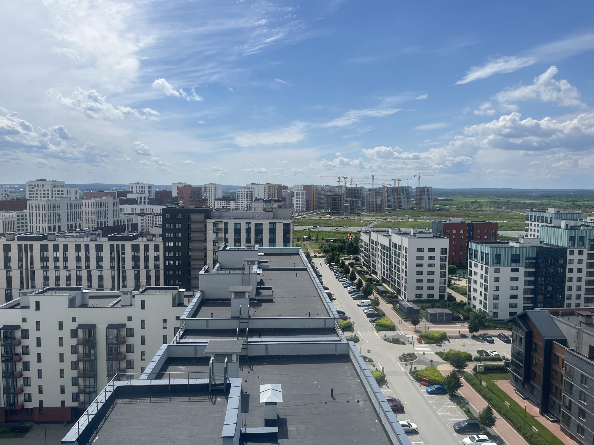 The image depicts a panoramic view of a modern urban district in Yekaterinburg, Russia, under a bright, partly cloudy sky. The foreground is dominated by the flat rooftops of several multi-story residential buildings, characterized by dark gray surfaces, parapets, and ventilation structures. In the mid-ground, a street is visible, lined with parked cars, pedestrian walkways with brick paving, and green spaces with scattered trees. The architecture of the residential buildings varies, with some featuring white facades and numerous windows, while others display brickwork and darker colored exteriors.
Further in the background, more residential complexes are under construction, indicated by the presence of several large tower cranes. Beyond the buildings, a vast expanse of green, undeveloped land stretches towards the horizon, suggesting the city's ongoing expansion into surrounding natural areas. The sky is a prominent feature, with scattered white cumulus clouds against a vibrant blue backdrop, indicating a clear day. The scene portrays a developing urban environment, a mix of completed and ongoing construction, with a clear emphasis on modern residential architecture and infrastructure.