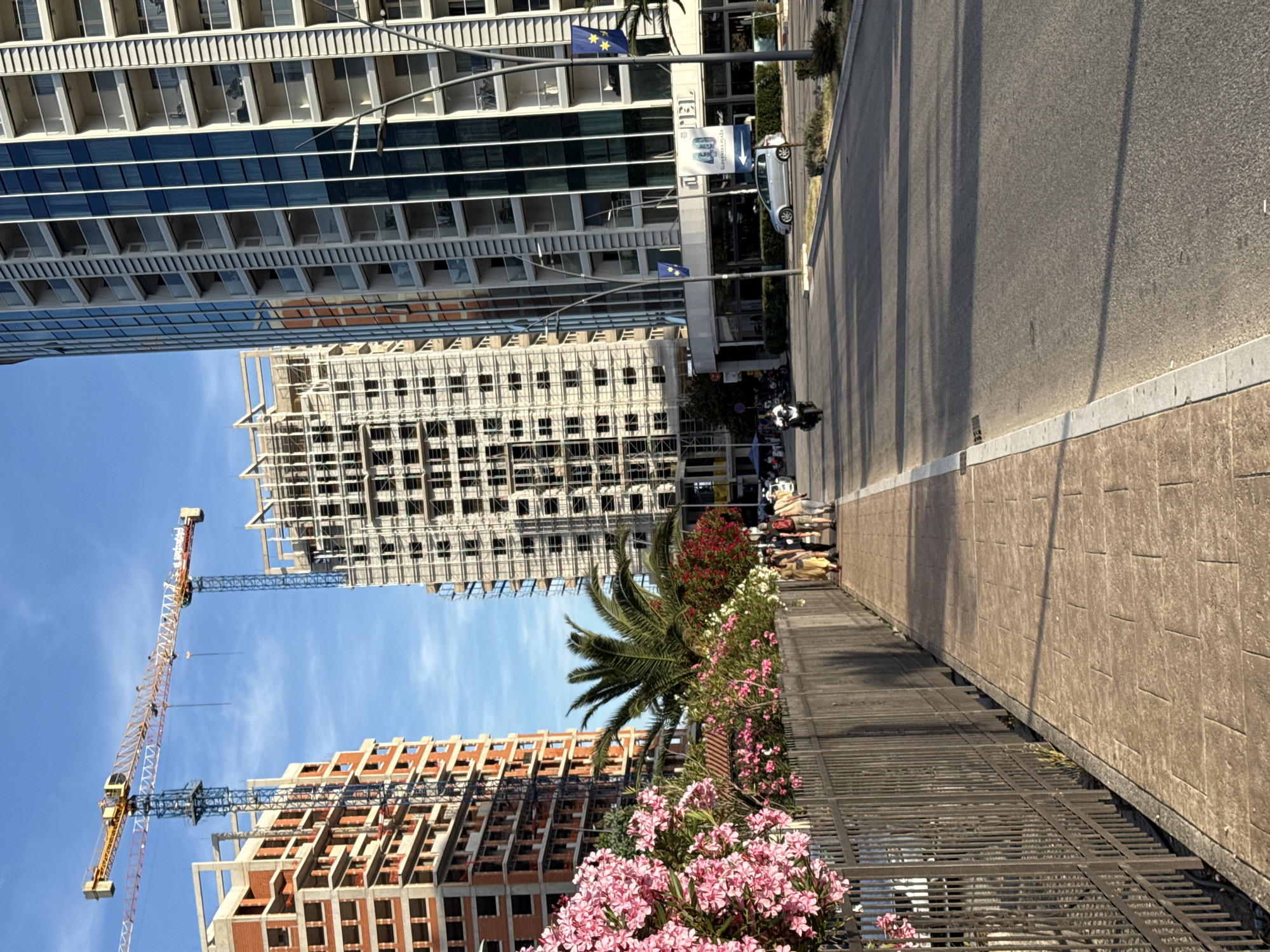 An urban street scene in what appears to be Budva, Montenegro, unfolds under a clear blue sky. On the left side of the image, a row of tall buildings dominates the skyline. The foremost is a modern, multi-story structure with numerous balconies and large, reflective glass windows, possibly a hotel, displaying European Union flags on poles attached to its facade. Partially visible text on this building reads "HOTEL" and a sign below shows "40" with an arrow and text that appears to be "Sajam turizma" (Tourism Fair) in a local script. Adjacent to this modern building, two other high-rise structures are actively under construction, characterized by exposed concrete frames, scaffolding, and some red brickwork. Two large tower cranes, one yellow and one blue, stand prominently against the sky, indicating ongoing development.
To the right, a paved road stretches into the distance, with a few vehicles visible. A silver car is parked or moving slowly, and a motorcycle with two riders is further down the street. Separating the road from a metal fence is a wide, textured sidewalk where several people are walking or standing. Along the length of the sidewalk, a vibrant strip of greenery features lush flowering bushes, predominantly pink oleander, alongside white and red blooms, and a tall palm tree. The time of day is bright, sunny daylight, evidenced by the clear sky and distinct shadows cast across the road. The overall impression is one of a busy, developing city.