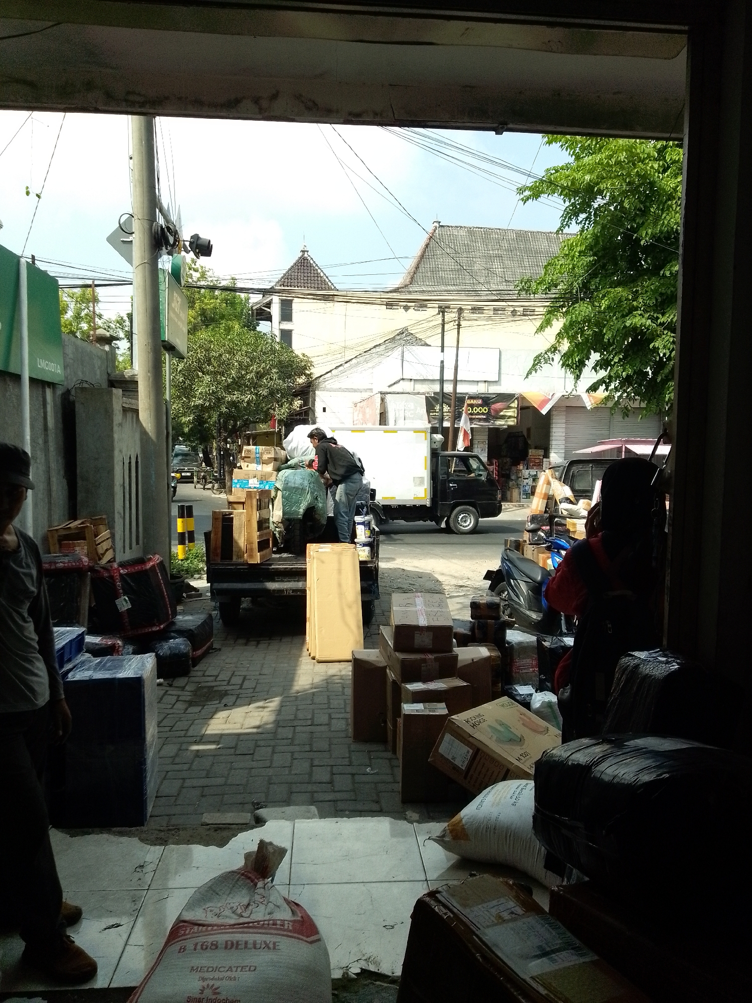 The image depicts an outdoor scene, likely a loading or unloading area in Tumenggungan, Indonesia, in front of a building with an open doorway. Several people are engaged in activities related to cargo. A person in a dark shirt and jeans is seen in the mid-ground, leaning over the back of a black pickup truck that is loaded with various boxes and items, some covered with green tarpaulin. Another person in a black cap and grey shirt is partially visible in the foreground on the left, facing away from the camera.
The ground is paved with interlocking bricks. Piles of boxes, some wooden crates, and wrapped packages are scattered around the area, both on the ground and on the truck. A motorcycle is parked behind the pickup truck. In the background, there are buildings with tiled roofs, trees, and utility poles with numerous wires. The sky is visible with some clouds. The lighting suggests it is daytime, with shadows cast on the ground.