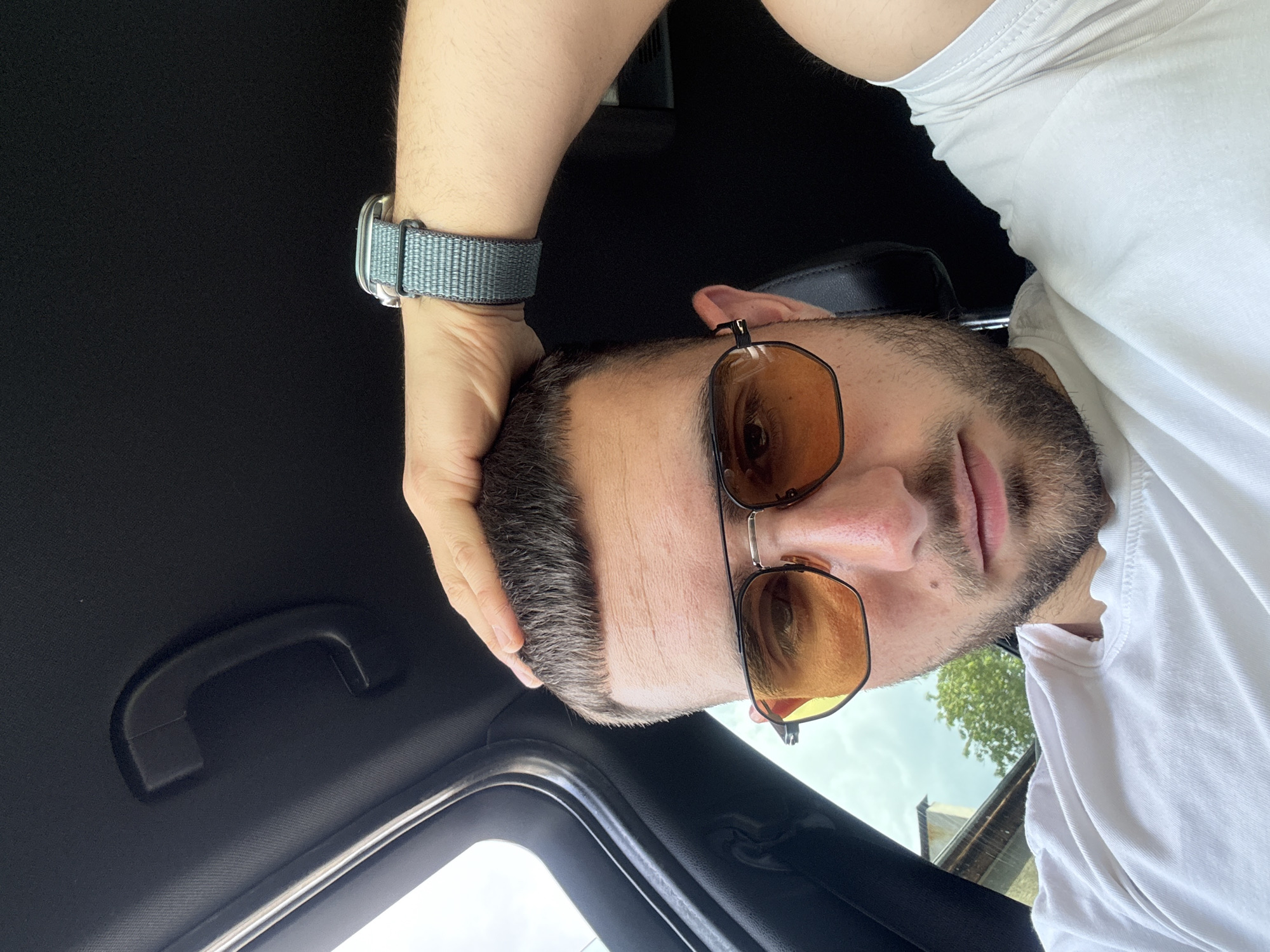 A selfie shows a man indoors, likely in a car, with a black interior roof and a grab handle. He is wearing a white t-shirt, sunglasses with amber lenses, and a light grey watch with a nylon strap. The man's hand rests on his head, and he is looking toward the camera. The background outside the car window appears to show greenery and buildings. The photo was not taken in Gabrovo, Bulgaria.