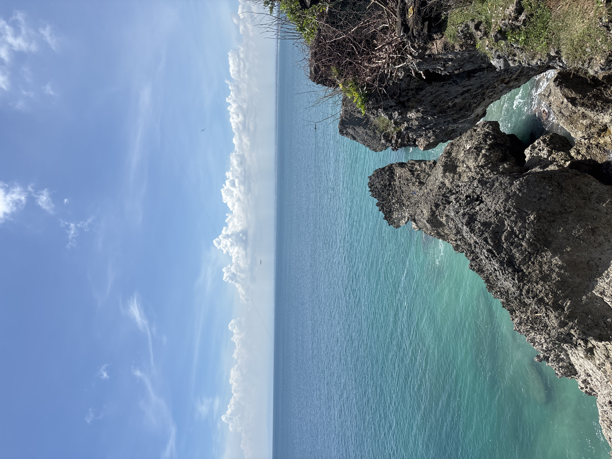 The image depicts a coastal scene in Unknown, Indonesia. Rocky cliffs with sparse vegetation overlook a vibrant, turquoise sea. The sky above is bright blue with streaks of cirrus clouds, and cumulus clouds line the horizon. There are objects in the sky that may be birds or airplanes.