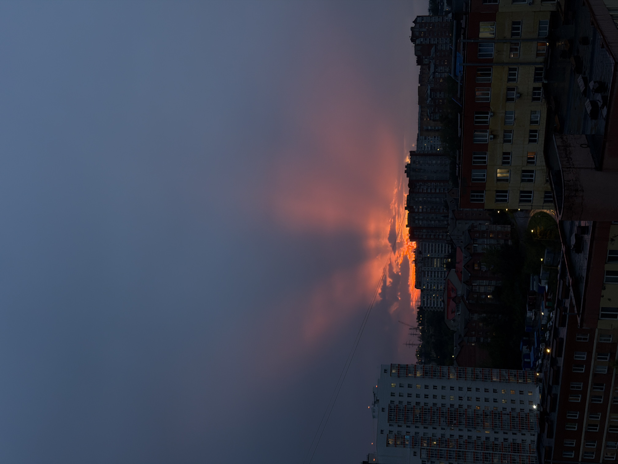 The image depicts a dramatic sunset over a cityscape, likely Krasnoyarsk, Russia. The sky is a deep blue transitioning to vibrant oranges and reds near the horizon, where the sun is setting behind clouds. Rays of light emanate upwards from behind a cluster of buildings, creating a striking visual effect. In the foreground and midground are apartment buildings of varying heights and architectural styles, characteristic of a Russian urban environment. Some buildings are brightly lit, suggesting the evening hours. The lighting is dim overall, with the sunset being the primary light source. Power lines and utility poles are visible, along with some trees. The perspective is from a higher vantage point, looking down onto the city. There are no people or animals clearly visible in the scene. The overall impression is one of natural beauty juxtaposed with urban development.