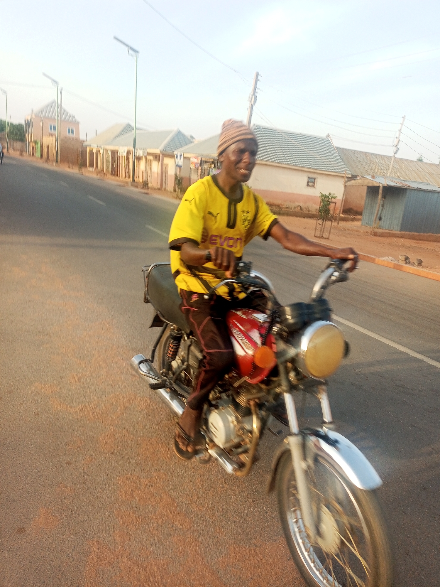 A man in a yellow shirt and striped hat rides a red motorcycle on a paved road in Tempure, Nigeria. He wears dark pants and sandals. The background features low buildings and utility poles. The sky is bright and clear.