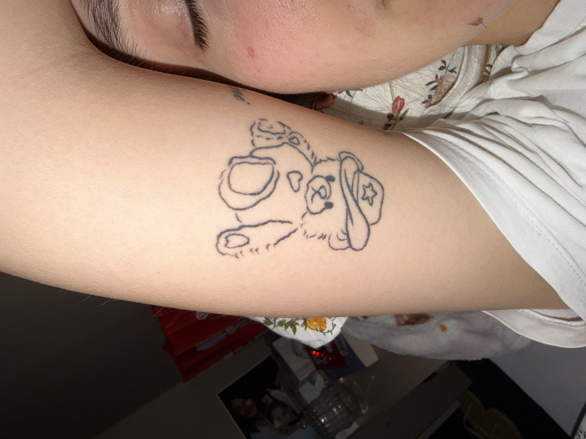 The media file presents a close-up, angled view of a person's upper body, focusing on their arm and a portion of their face. The primary subject is a person, likely a young adult, whose forehead, eyebrow, and a portion of their cheek are visible on the upper left side of the frame. Their arm, positioned diagonally across the center, features a prominent tattoo.
The tattoo, located on the upper arm, is a black outline drawing of a cartoon-style dog's head. The dog has large, round eyes, what appears to be a muzzle or snout, and is wearing a hat or bandana adorned with a star emblem on its left side. Fine hair is visible on the person's arm.
The person is wearing a white t-shirt, part of which is visible along the right side of the arm. Beneath the arm, a garment with a floral pattern, possibly part of a top or pajama, is partially visible.
The background is dark and mostly out of focus, suggesting an indoor setting with subdued lighting. Objects discernible in the blurred background include a red item (possibly a bag or container) on the lower left, a framed picture or screen showing two indistinct blurred faces, and a clear glass or container with liquid.
The activity appears to be the person resting or lying down, given the relaxed posture and partial view of their face. There is no visible text in the image. The time of day cannot be definitively determined, but the dim lighting suggests an evening or nighttime setting.