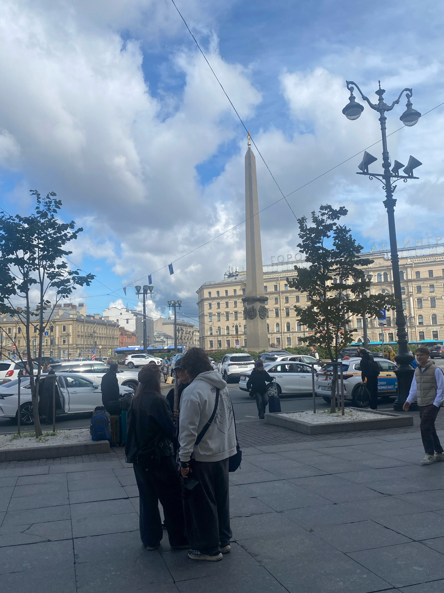 In Saint Petersburg, Russia, an obelisk stands tall against a cloudy sky, a landmark likely in Palace Square given the surrounding architecture. Cars are parked and moving on a street in front of a light-colored, multi-story building with numerous windows. Several trees are visible, some planted in square planters with sand. In the foreground, a group of four individuals stands on a paved walkway. Two are facing each other and conversing, while the other two are positioned slightly to the side. One person is wearing a white hooded sweatshirt and dark pants, another a black jacket and dark pants, and the third a dark jacket and black pants with a hat. The fourth person, to the right, is a young male in a light-colored vest and brown pants. A lamppost with multiple lights is prominent on the right side of the frame. The overall scene depicts a busy urban environment with a mix of historical and modern elements.