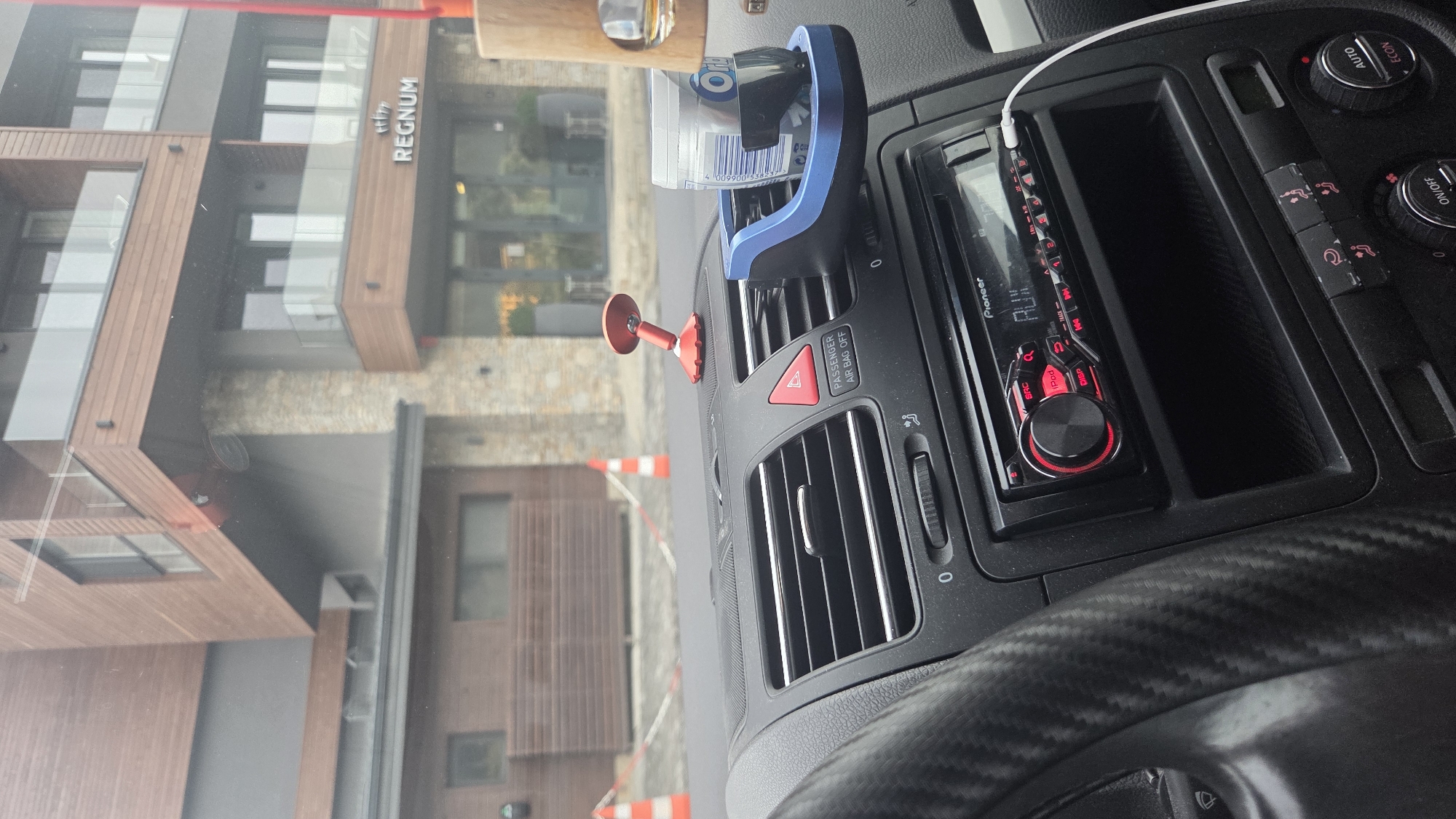 This is a close-up interior shot of a car's dashboard, with a modern building visible through the windshield. The dashboard features a Pioneer car stereo with red lighting, air vents, and a cup holder attached to one of the vents. The cup holder contains a cylindrical object, possibly a container of gum or mints, and a wooden-framed object hangs from the windshield, holding a glass with amber liquid. A red suction-cup phone mount is also visible on the dashboard. The exterior view shows a building with wooden and stone facade, balconies with glass railings, and the prominent sign "REGNUM". Orange traffic cones and red and white tape are visible in the background, suggesting a cordoned-off area. The lighting indicates it is daytime, but overcast.