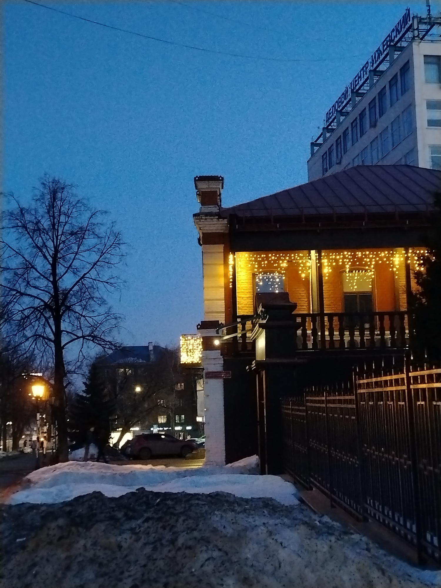 The image shows a street scene in Izhevsk, Russia at dusk. Snow is piled along the roadside. A wrought-iron fence runs along the right side of the frame. A two-story brick building is decorated with illuminated string lights. A business center with a sign in Russian "ДЕЛОВОЙ ЦЕНТР ИЖЕВСКИЙ" (Izhevsk Business Center) is visible in the background. A bare tree stands on the left side of the frame next to a street lamp. A car and other buildings are visible in the background.