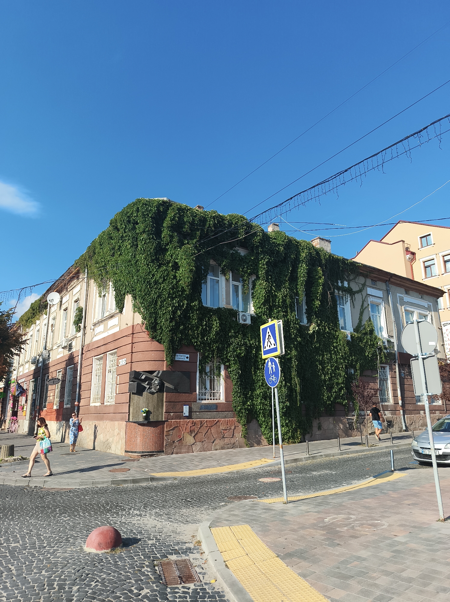 A street corner in Ternopil, Ukraine, features a multi-story building covered in lush green ivy. The building has a pinkish-brown facade with white trim around the windows. A monument, possibly a bust on a stone pedestal, is located on the corner of the building. Across the street, there is a crosswalk with pedestrian and bicycle signs. The street is paved with cobblestones, and a silver car is parked on the right side of the road. Several people are visible walking on the sidewalks and crossing the street. The sky is a clear, bright blue, suggesting a sunny day.