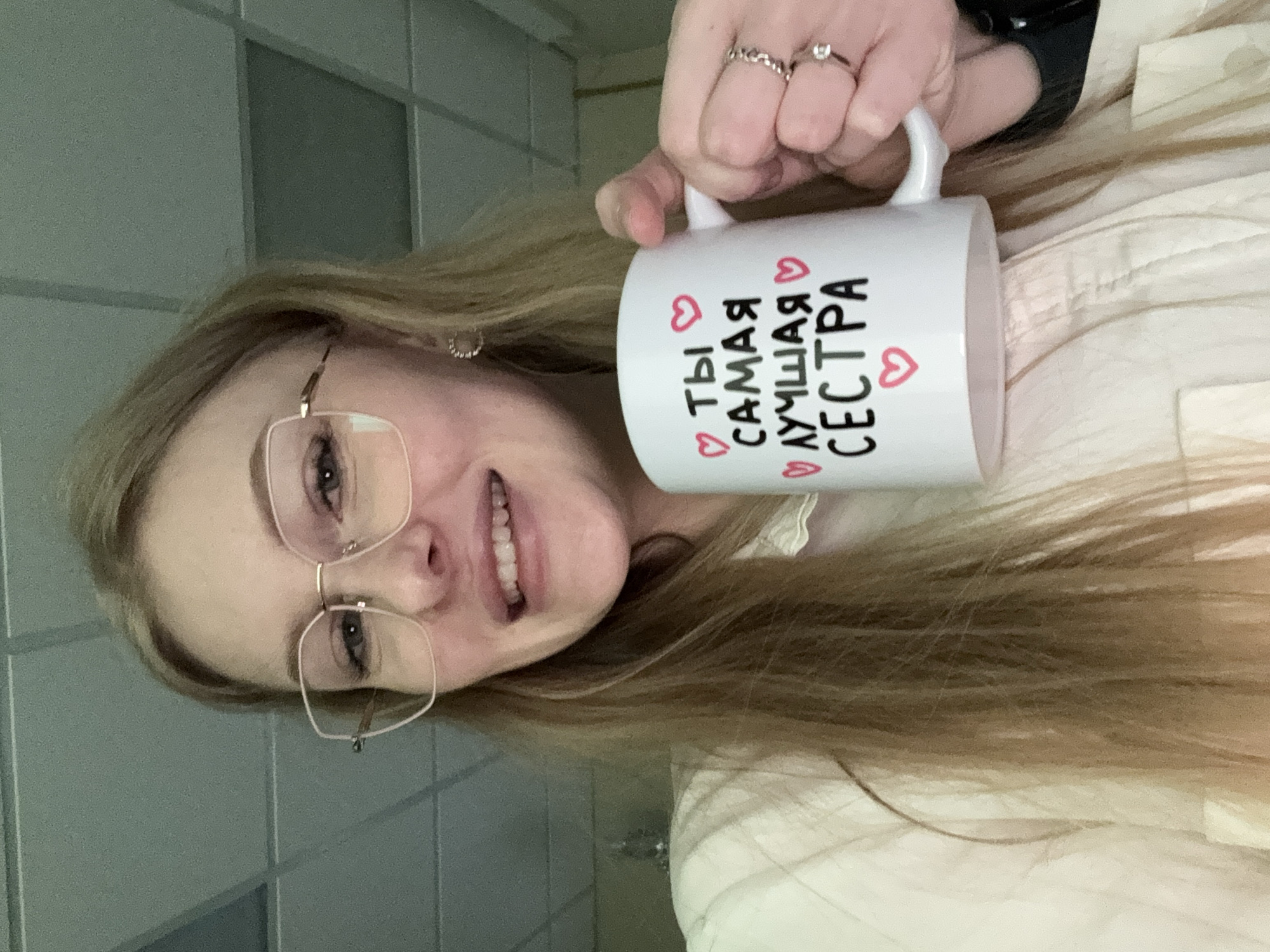 An indoor portrait features a young woman with long, light brown hair and clear-rimmed glasses, smiling broadly at the camera. She holds a white ceramic mug in her right hand, which is adorned with two rings on her fingers and a partially visible watch on her wrist. The mug displays Russian text in black, interspersed with small red heart symbols. The text reads "ТЫ САМАЯ ЛУЧШАЯ СЕСТРА," translating to "YOU ARE THE BEST SISTER." The woman wears light-colored clothing. The background consists of a light-toned wall with subtle grid patterns, suggesting an indoor setting with bright, even lighting. The image conveys a cheerful and personal sentiment, likely taken in Perm, Russia.