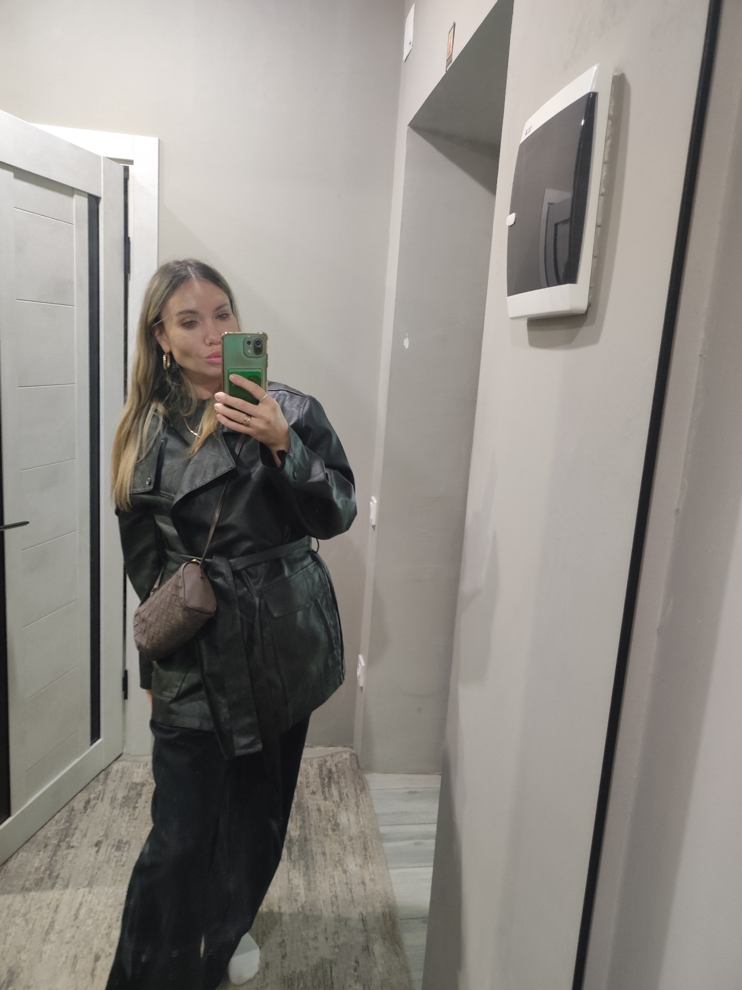 A woman is taking a selfie in what appears to be the hallway of an apartment in Votkinsk, Russia. She is wearing a black leather jacket, black pants, and white sneakers, and carries a brown woven handbag. The hallway has light gray walls, a white door, and wood-patterned flooring. An electrical panel is mounted on the wall next to a doorway.