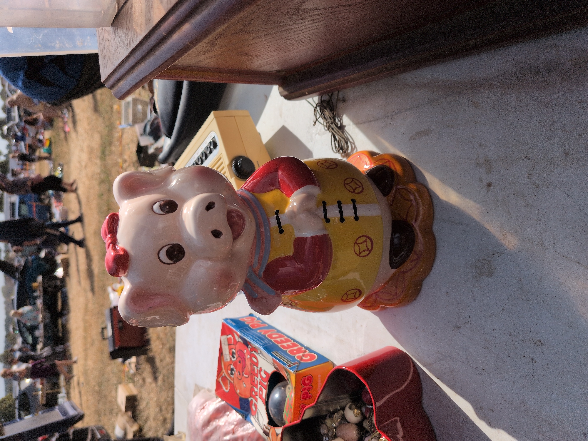 This is a medium shot, eye-level, outdoor photograph taken at a flea market in Claverley, United Kingdom. The focus is on a ceramic piggy bank in the foreground, positioned on a white, textured surface. The piggy bank is predominantly pink with a red bow on its head and is wearing a yellow tunic with red trim and black detailing. It is standing on an orange base. To the left of the piggy bank is a red container filled with assorted items, appearing to be jewelry or trinkets. Behind the red container is a blue and orange box with the text "CREEDY PIG" visible. A small, yellow electronic device with buttons and a dial is behind the piggy bank. The background is blurred, showing a grassy field with several people browsing at stalls. The overall impression is of a typical outdoor market scene under a bright sky.