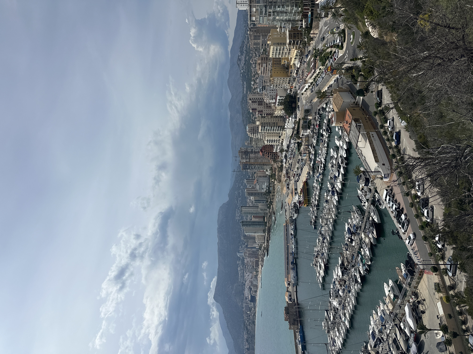 The image shows a harbor densely packed with boats and yachts, situated along the coast of a city. Buildings, some tall and modern, line the waterfront behind the harbor. Further back, a mountain range is visible. The sky is partially cloudy. Roads and parked cars can be seen along the harbor's edge. A line of trees borders the harbor. The location may be San Gabriel, Spain.